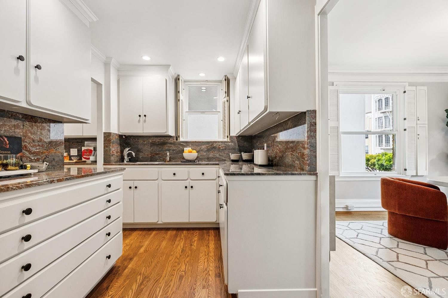 This is a well-lit kitchen with white cabinetry and dark countertops. The kitchen features a window above the sink and hardwood floors. The style is traditional and clean, creating a welcoming atmosphere.