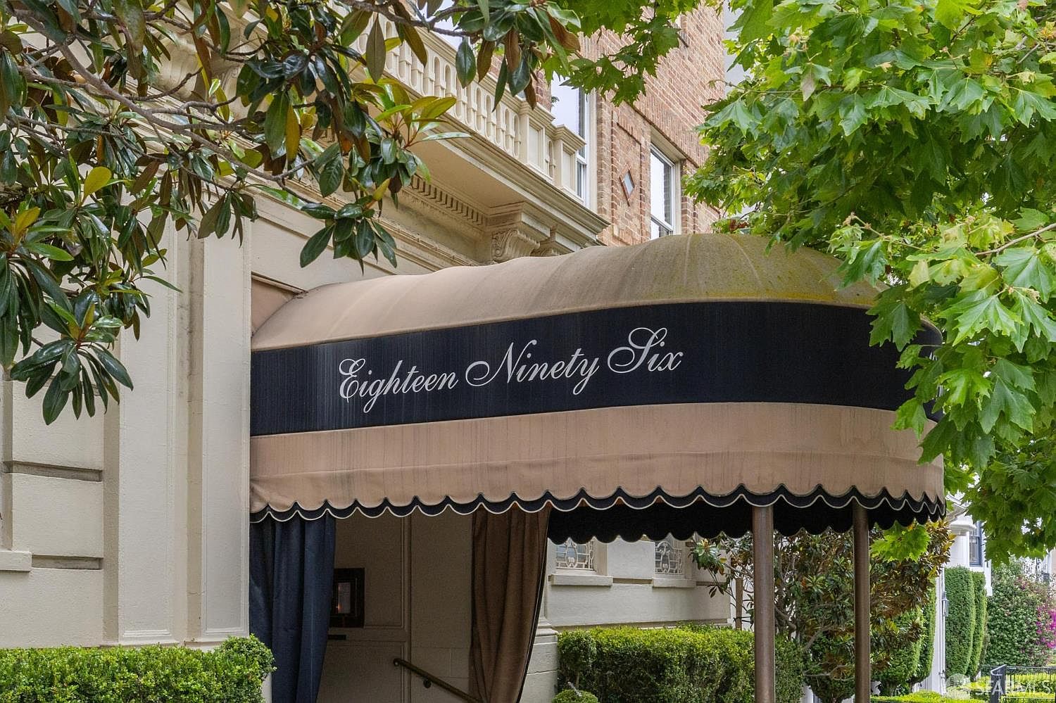 The image showcases the elegant entryway of a building, featuring a prominent awning with the address "Eighteen Ninety Six" displayed in a classic font. The awning is supported by pillars and framed by lush greenery, creating a welcoming and sophisticated entrance. The building's facade exhibits traditional architectural details, suggesting a well-maintained and stately property.