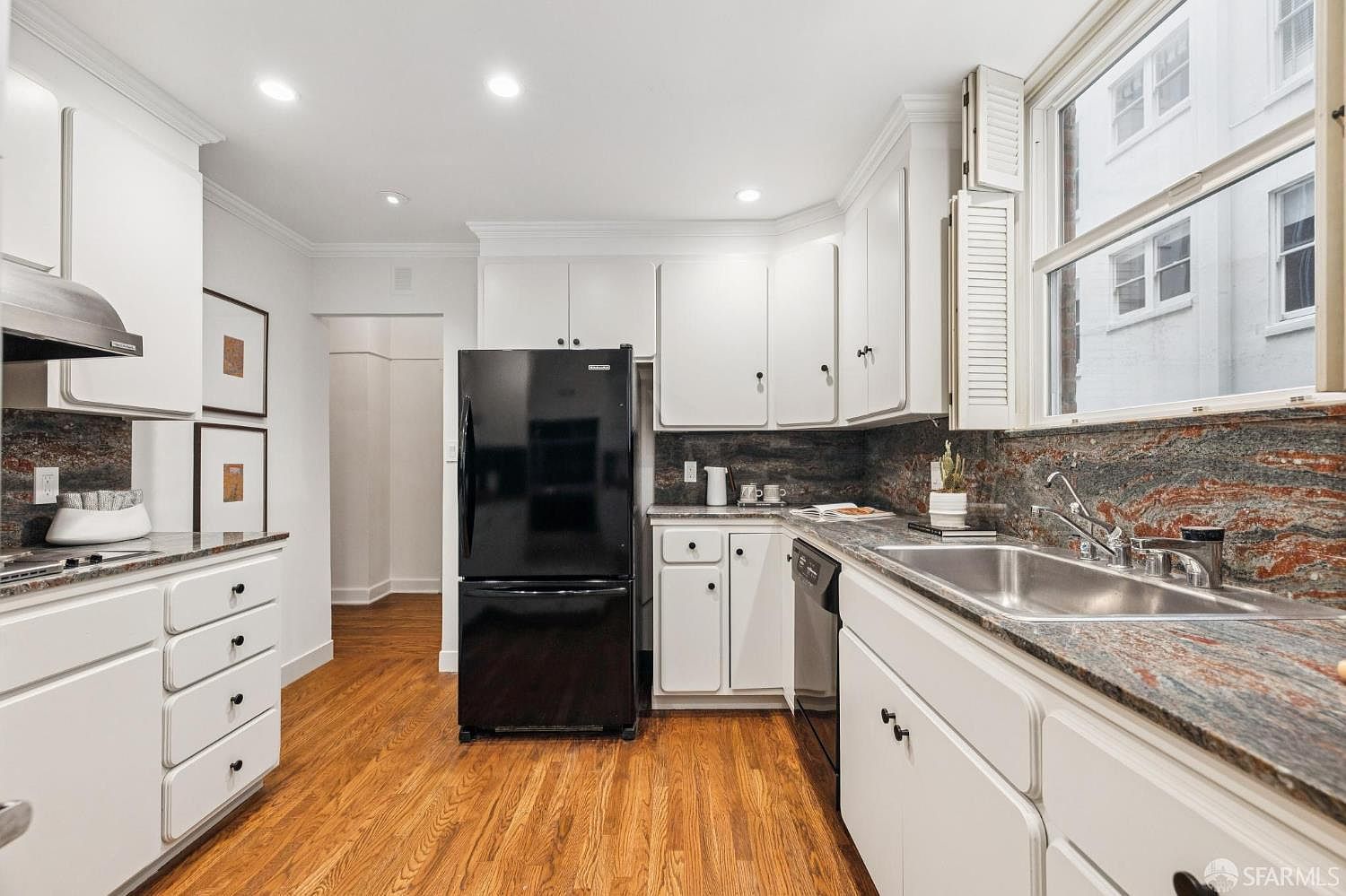 This is a well-lit kitchen featuring white cabinetry, a black refrigerator, and granite countertops with a unique reddish-brown veining pattern. The hardwood floors add warmth to the space, while the stainless steel sink and faucet provide a modern touch. The window above the sink offers natural light and a view of the exterior.