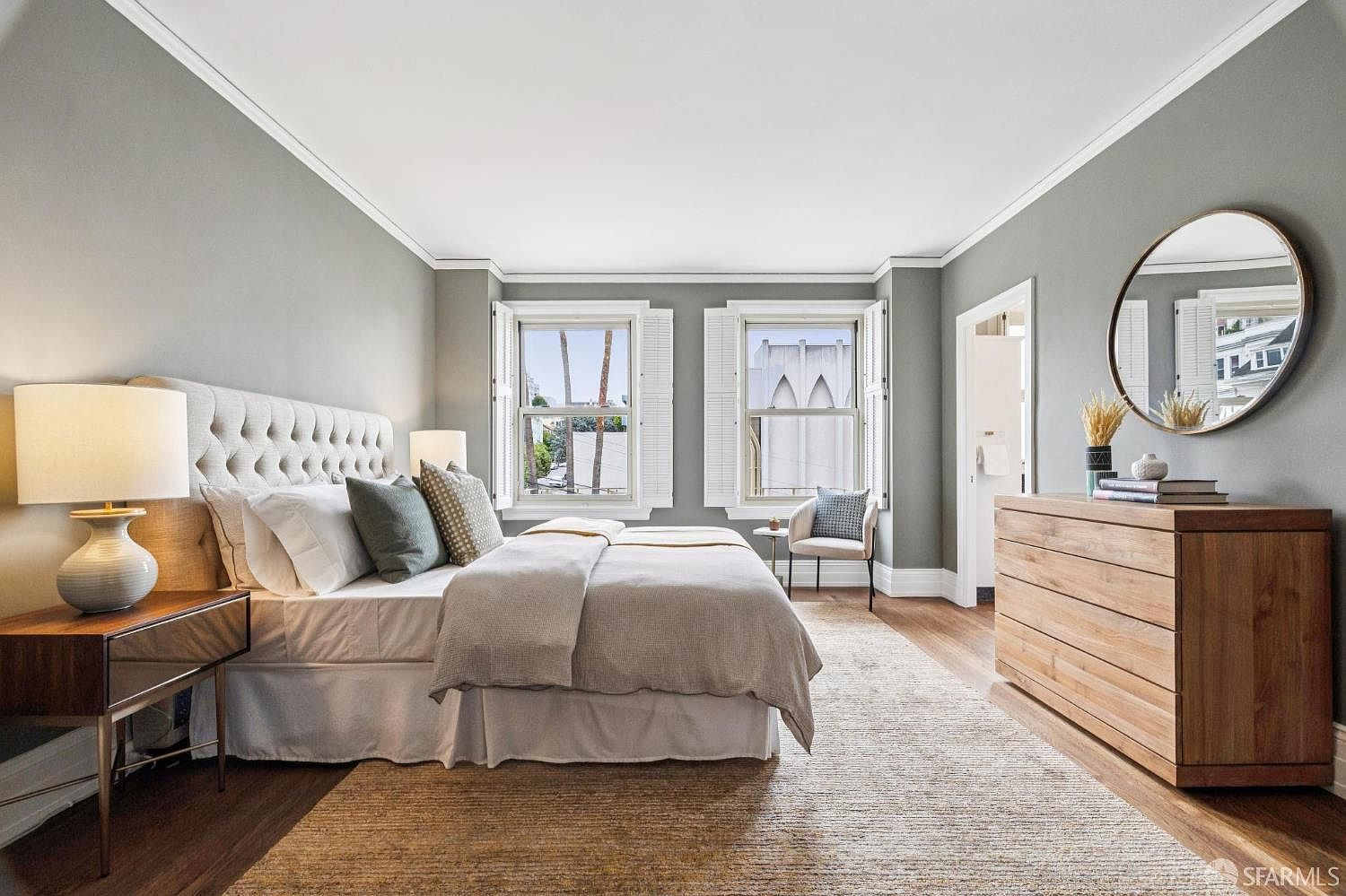 This is a well-lit primary bedroom featuring a tufted headboard bed with neutral bedding, complemented by a wooden nightstand and dresser. Two windows with white shutters provide natural light, and a round mirror hangs above the dresser. The room is decorated in a neutral color palette with hardwood floors and a large area rug, creating a serene and inviting atmosphere.