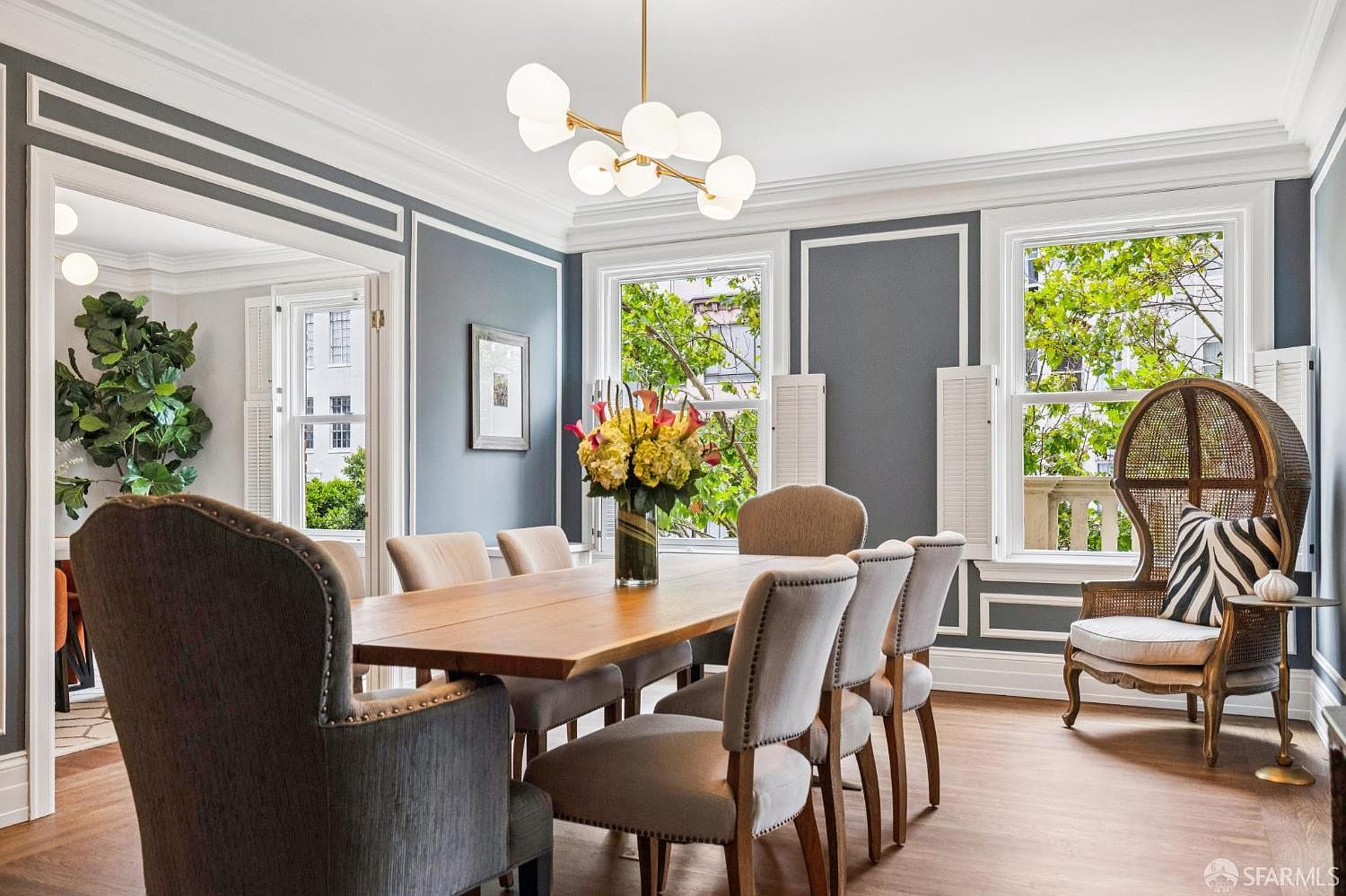This is a dining room featuring a large wooden table surrounded by upholstered chairs. A modern chandelier hangs above the table, and natural light streams in through the windows, which are framed by white shutters. The walls are painted a muted gray with white trim, creating a sophisticated and inviting atmosphere.