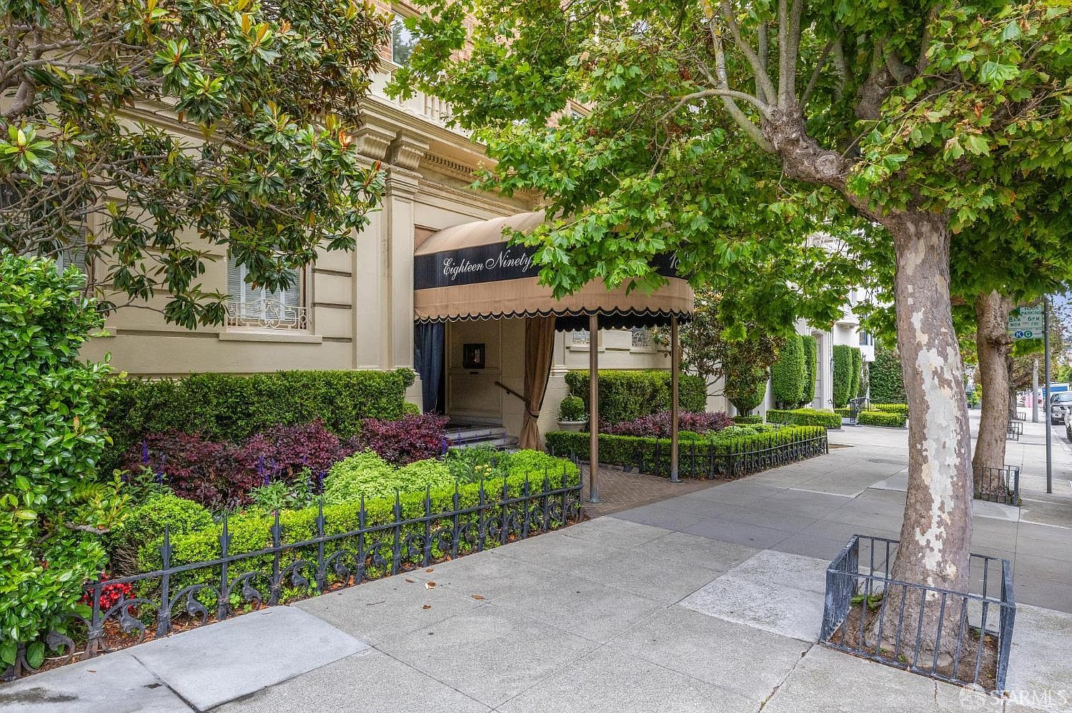 The image showcases the elegant entryway of a building, featuring a tan awning with black trim and the address "Eighteen Ninety" prominently displayed. Lush, manicured landscaping with a variety of plants and a decorative black fence add to the curb appeal. The building's facade is a light beige color, and the overall impression is one of sophistication and refined taste.