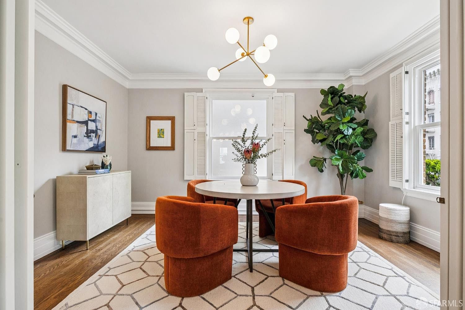 This is a well-lit dining room featuring a round white table surrounded by four orange velvet chairs. A modern chandelier hangs above the table, and a large fiddle-leaf fig tree adds a touch of nature to the space. The room is decorated with artwork and a stylish sideboard, creating a sophisticated and inviting atmosphere.