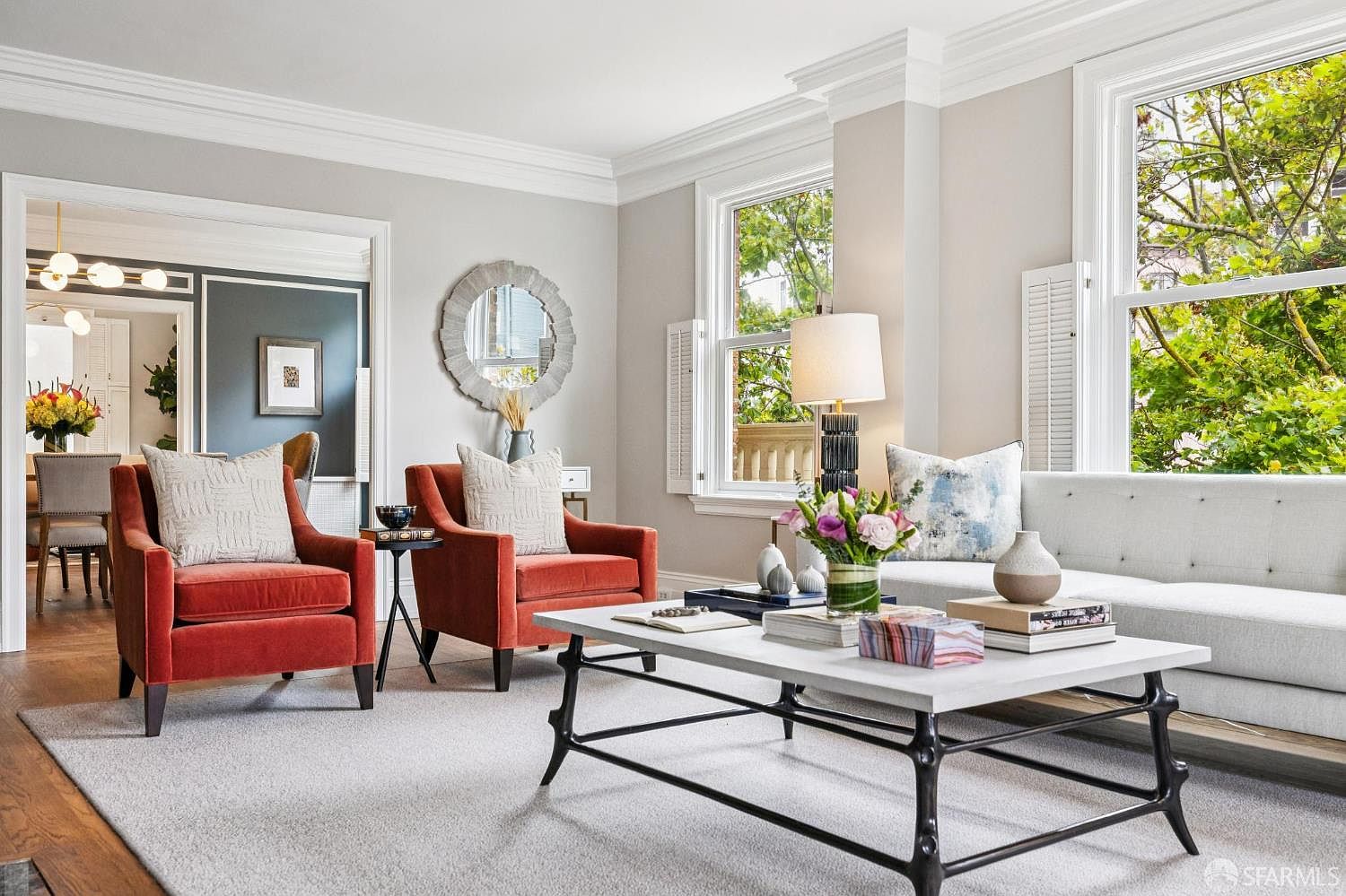 This is a well-lit living room featuring two rust-colored armchairs with patterned pillows, a light gray sofa, and a white coffee table with a black metal base. The room has large windows with white shutters, allowing natural light to illuminate the space. A decorative mirror hangs on the wall, and an open doorway leads to another room, creating a sense of spaciousness.