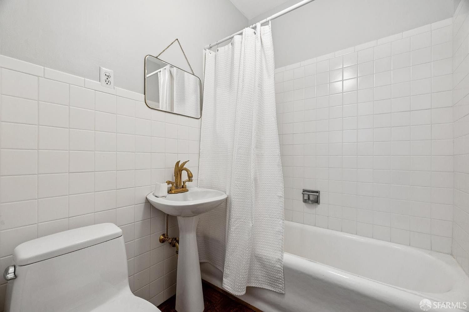 This is a bathroom featuring white tiled walls, a pedestal sink with a gold-colored faucet, and a toilet. A framed mirror hangs above the sink, and a white shower curtain is drawn across the bathtub. The bathroom has a clean and simple design.