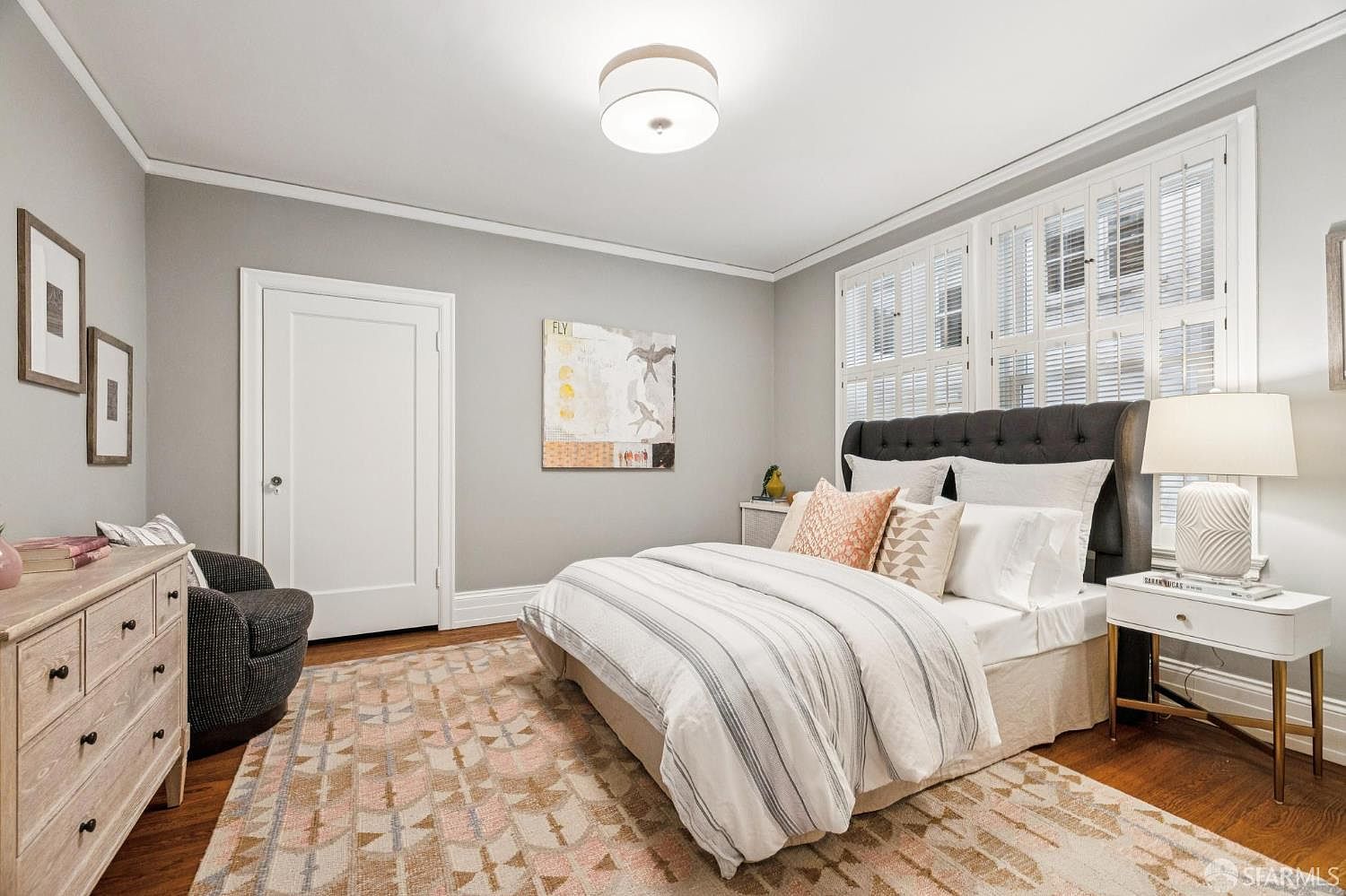 This is a well-lit primary bedroom featuring a bed with a tufted headboard, striped bedding, and decorative pillows. A white nightstand with a lamp sits beside the bed, and a patterned rug covers the hardwood floor. The room is painted in a neutral gray tone, complemented by white trim and shutters on the windows.