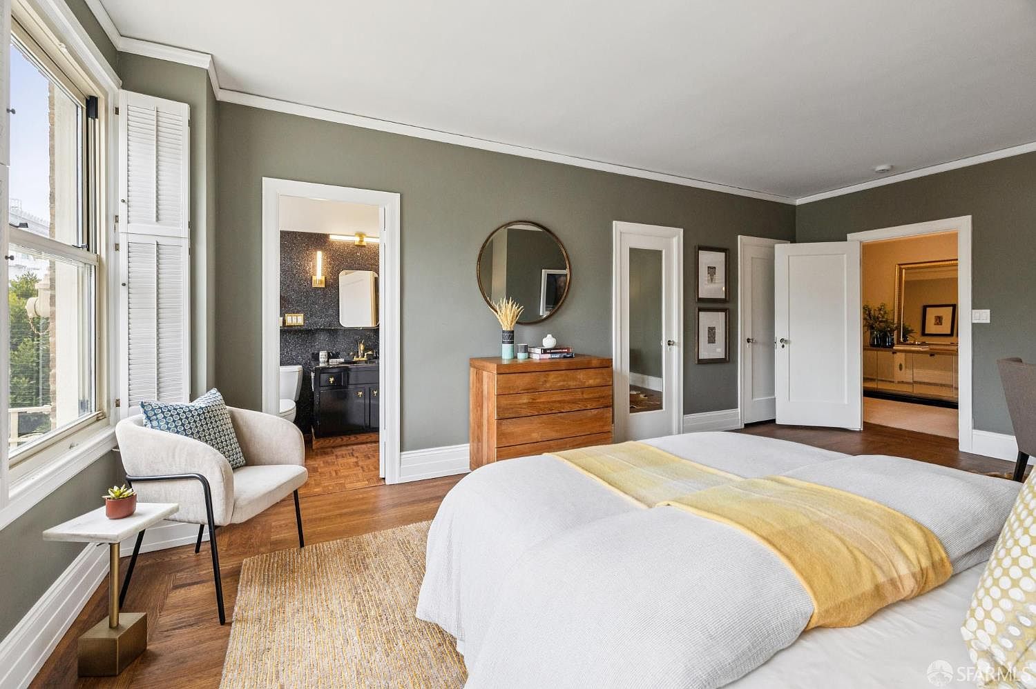 This is a primary bedroom featuring a large bed with white linens and a yellow throw, a wooden dresser with a round mirror above it, and a comfortable armchair by the window. The walls are painted in a muted green tone, complemented by white trim and doors. The room has a warm and inviting atmosphere, with hardwood floors and a view into an adjacent bathroom and hallway.