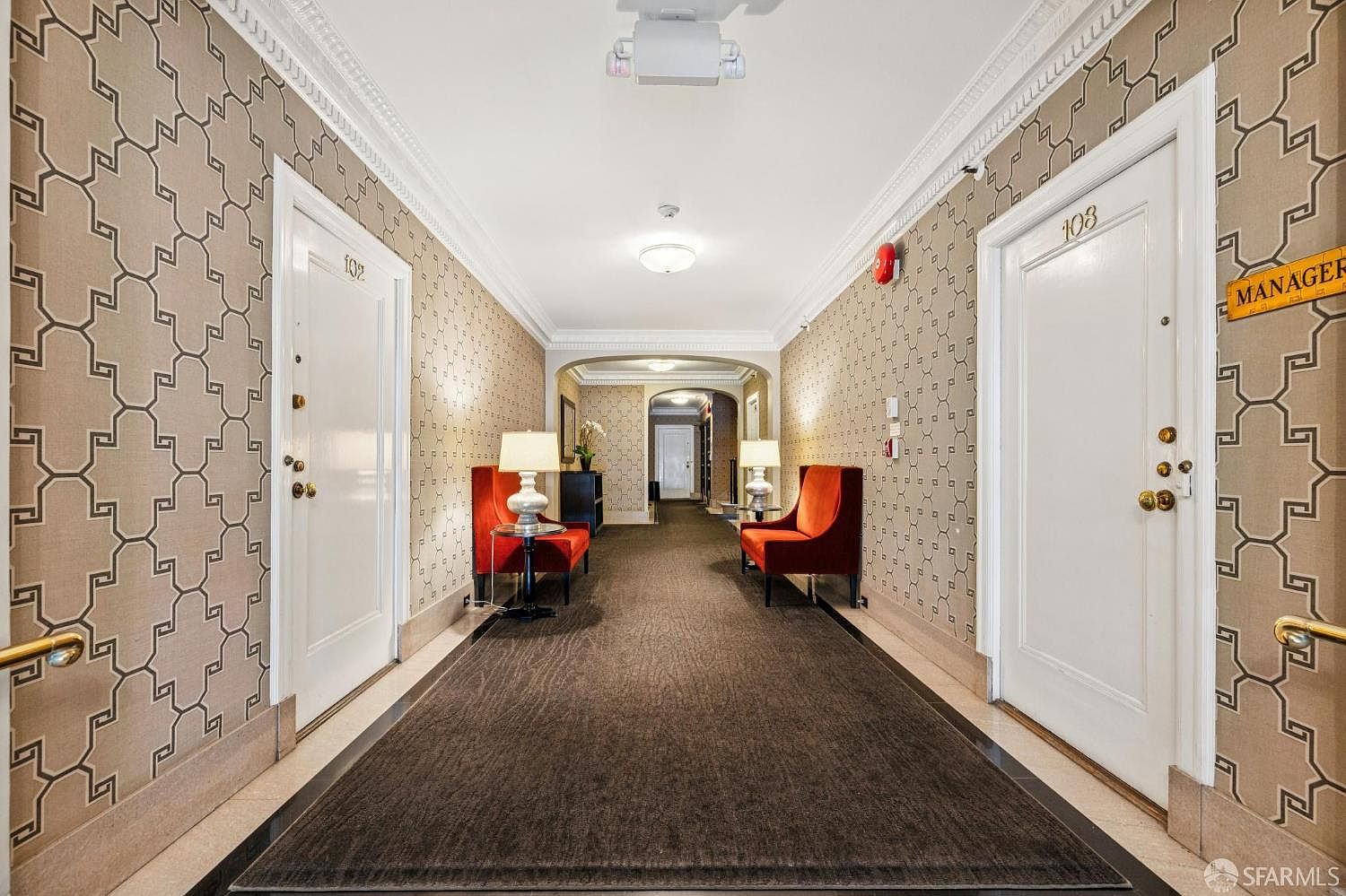 This is an interior shot of a well-maintained hallway in an apartment building. The hallway features patterned wallpaper, white doors with unit numbers, and elegant seating areas with red chairs and side tables. A long runner rug stretches down the center of the hallway, adding a touch of sophistication.