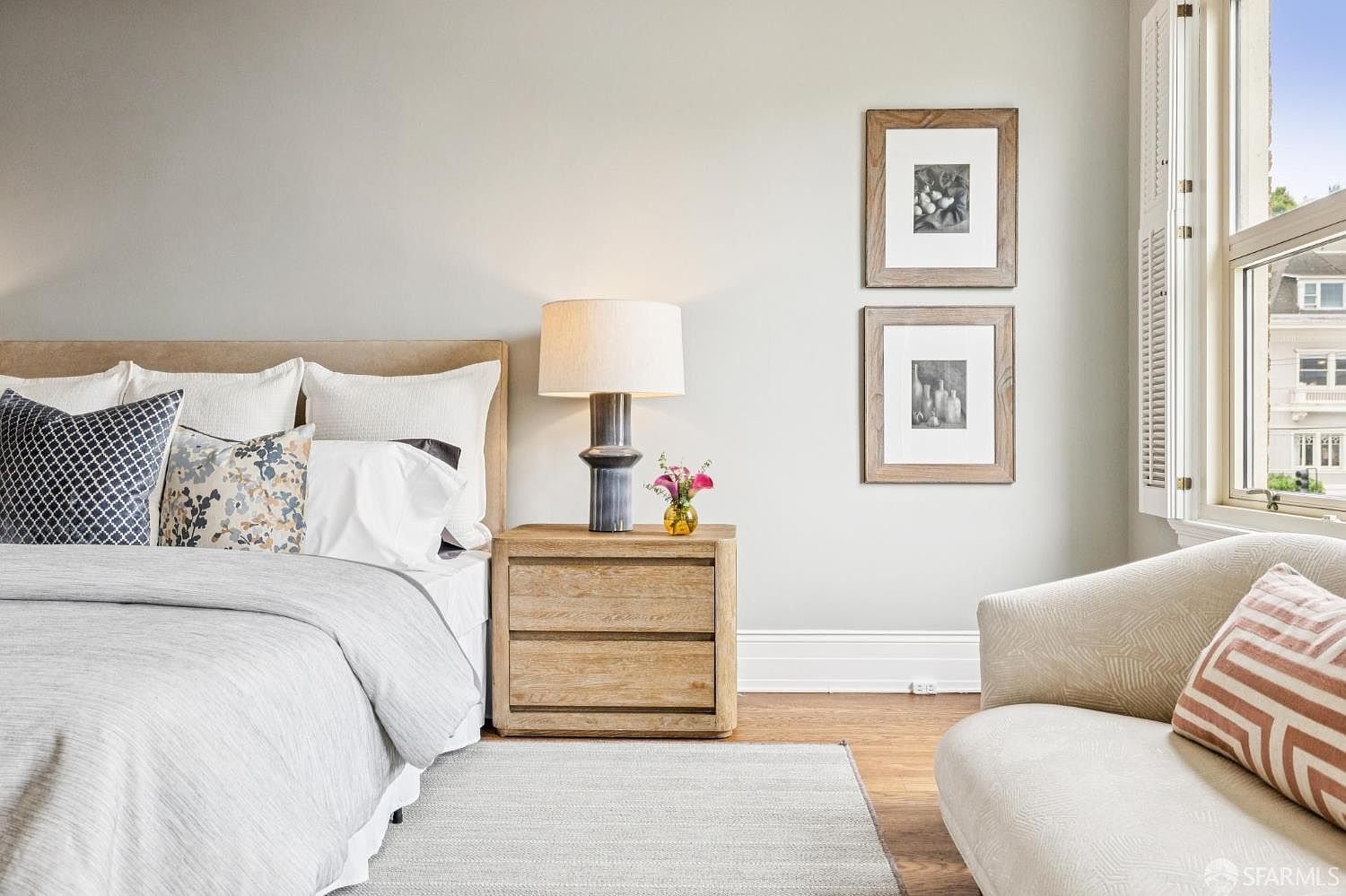 This is a well-staged primary bedroom featuring a neutral color palette. The bed is neatly made with decorative pillows and a light-colored duvet. A wooden nightstand with a lamp and a small vase of flowers sits beside the bed, and framed artwork hangs on the wall above a cozy armchair, creating a serene and inviting atmosphere.