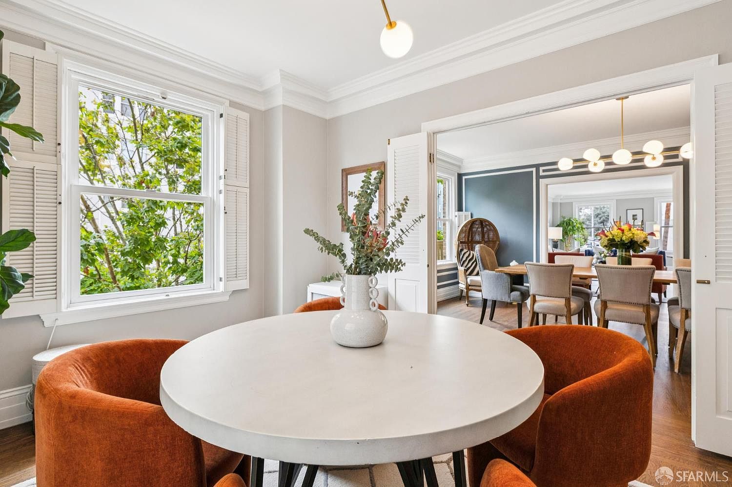 This is an interior shot of a dining room featuring a round table with orange chairs. The room is well-lit with natural light coming through a large window. An open doorway leads to another dining area, creating a sense of spaciousness and flow.
