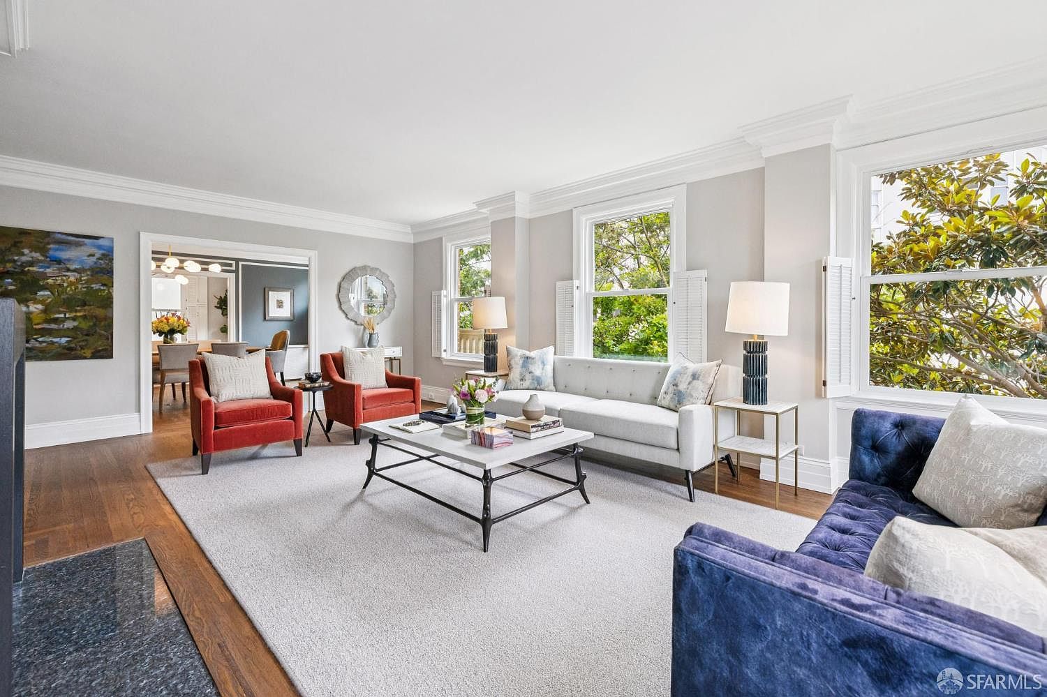This is a well-lit living room featuring a neutral color palette with pops of color from the red armchairs and blue sofa. The room is furnished with a white sofa, a marble-topped coffee table, and side tables with lamps. Large windows provide natural light and views of the outdoors, enhancing the room's spacious feel.