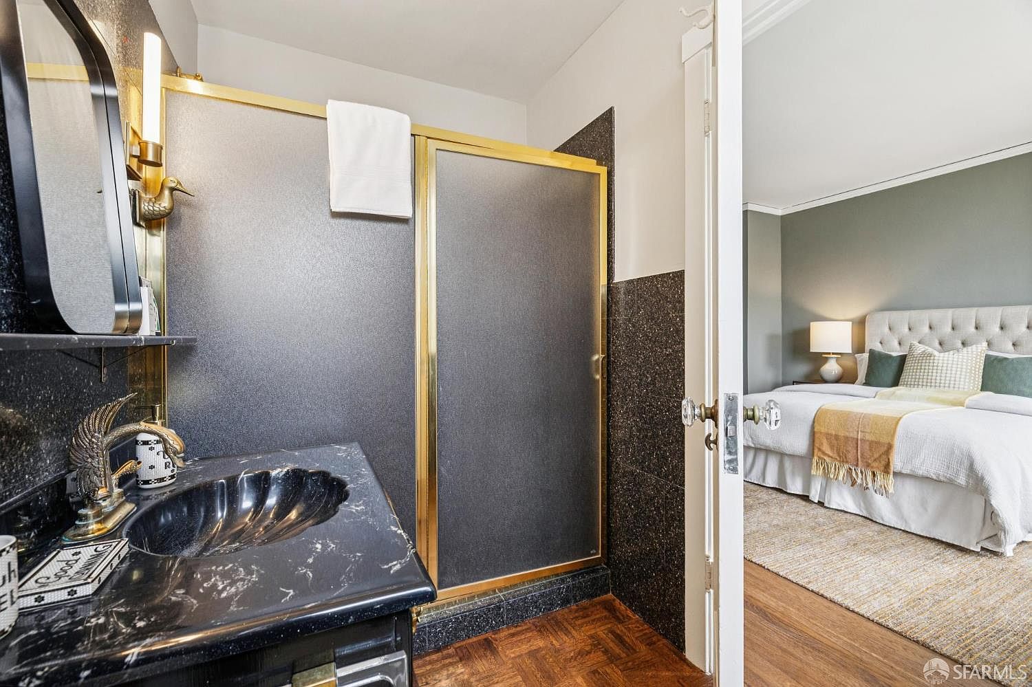 This is a primary bathroom featuring a unique black marble sink and countertop, complemented by gold fixtures and a shower with a frosted glass door. The bathroom has a vintage aesthetic, with dark speckled tiles on the wall and a herringbone wood floor. An open doorway leads into a bedroom with a tufted headboard and neutral decor.