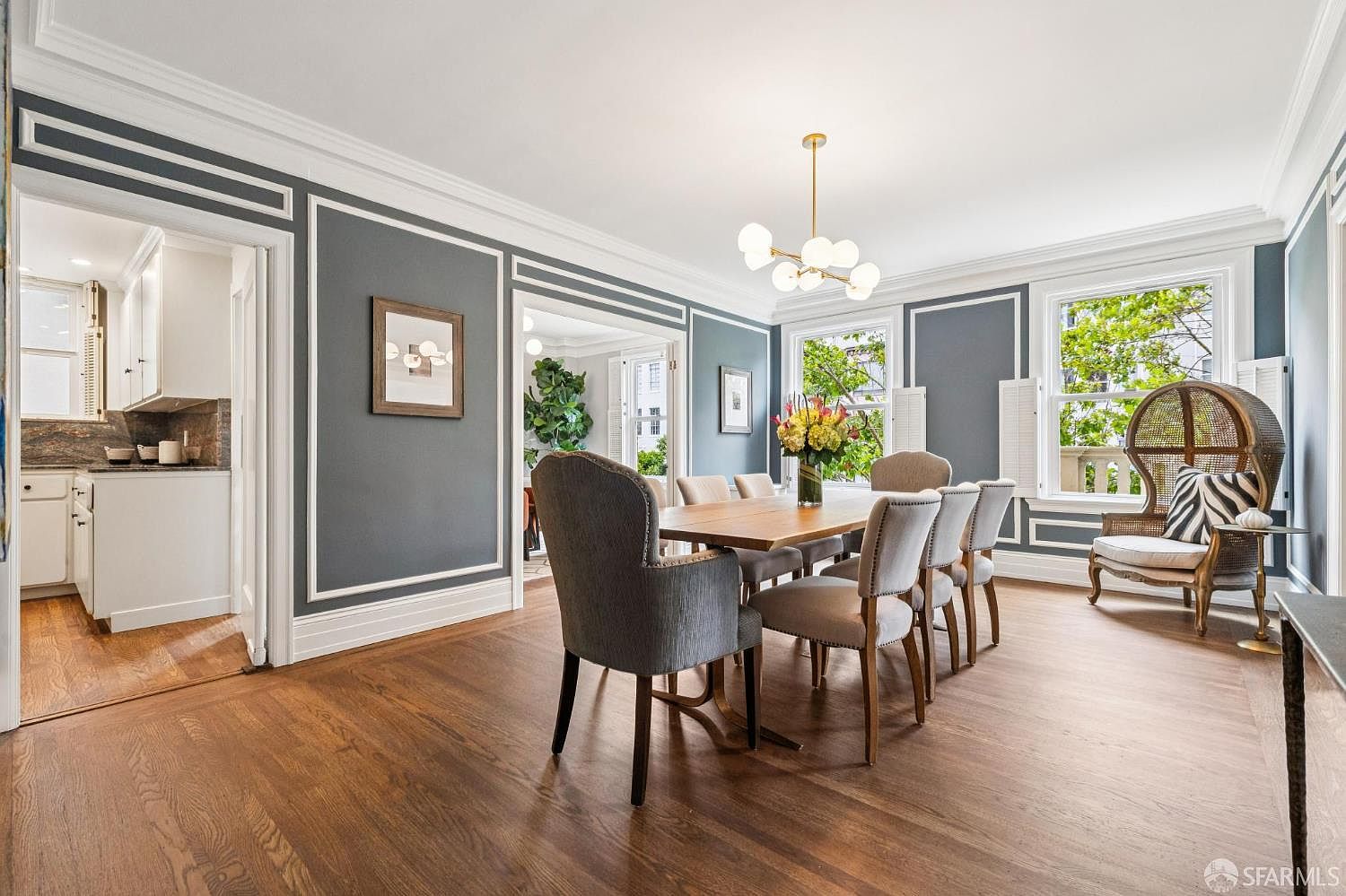 This is an interior shot of a dining room featuring a long wooden table surrounded by upholstered chairs. The walls are painted a dark gray with white trim, and a modern chandelier hangs above the table. Natural light streams in through the windows, highlighting the hardwood floors and creating a warm, inviting atmosphere.