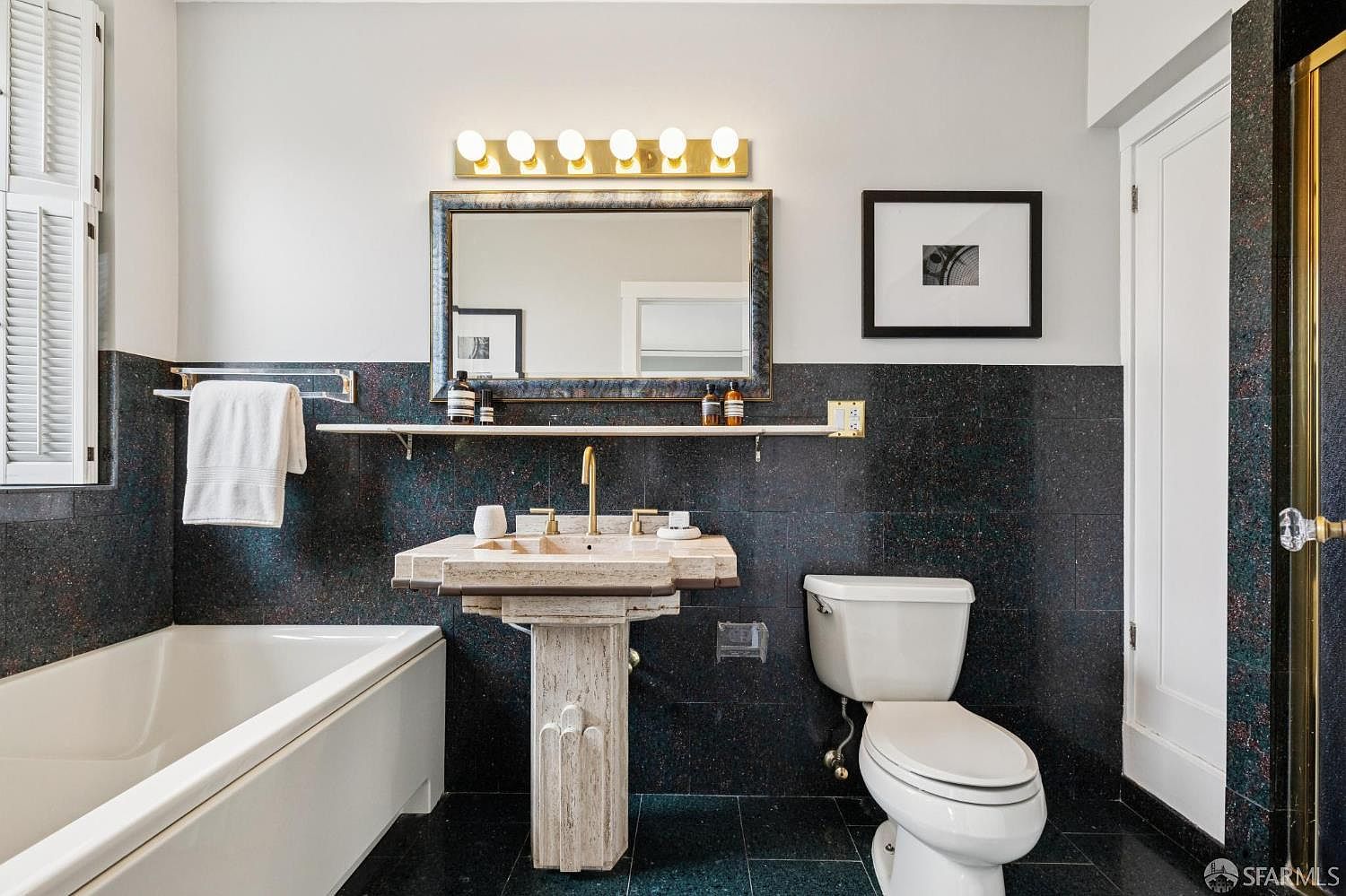 This is a bathroom featuring a unique stone sink and pedestal, a toilet, and a bathtub. The walls are tiled in a dark, speckled pattern, contrasting with the white fixtures and upper wall. A framed mirror with a light fixture above it adds to the room's character, while a framed picture hangs on the wall.