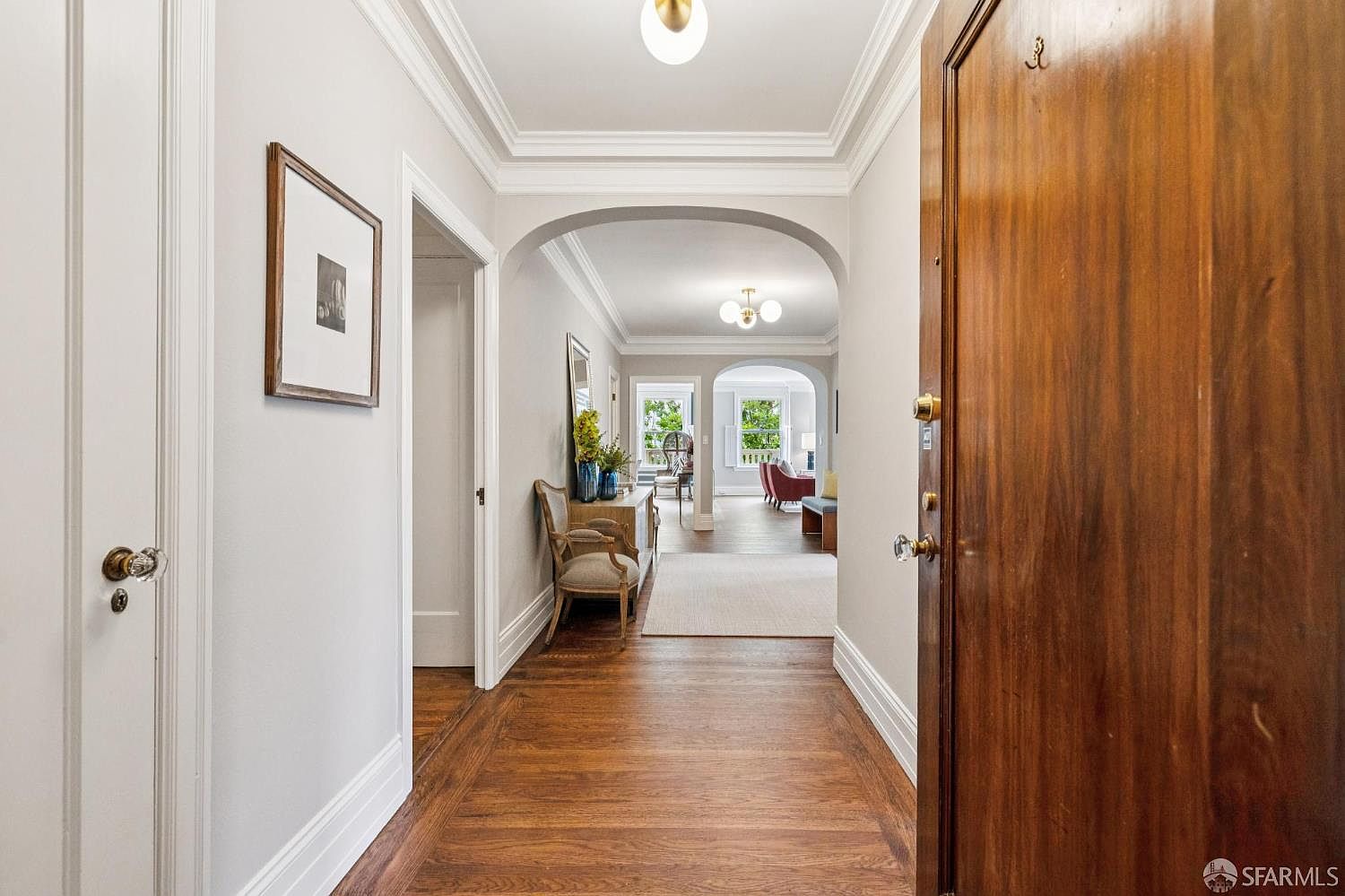 This interior shot showcases a grand hallway with rich hardwood flooring and elegant architectural details, including arched doorways and crown molding. A glimpse into the living spaces reveals a comfortable and inviting atmosphere, while a partially open wooden door adds depth and perspective to the scene. The overall impression is one of classic sophistication and timeless appeal.