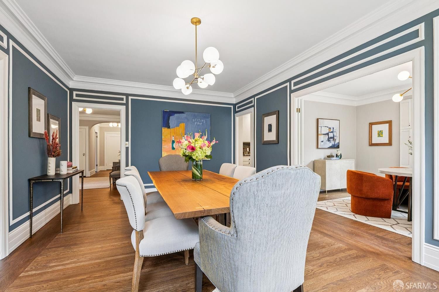 This is a beautifully appointed dining room featuring a large wooden table surrounded by upholstered chairs. The walls are painted a deep blue with white trim, and a modern chandelier hangs above the table. Artwork adorns the walls, and the hardwood floors are laid in a herringbone pattern, creating a sophisticated and inviting space.