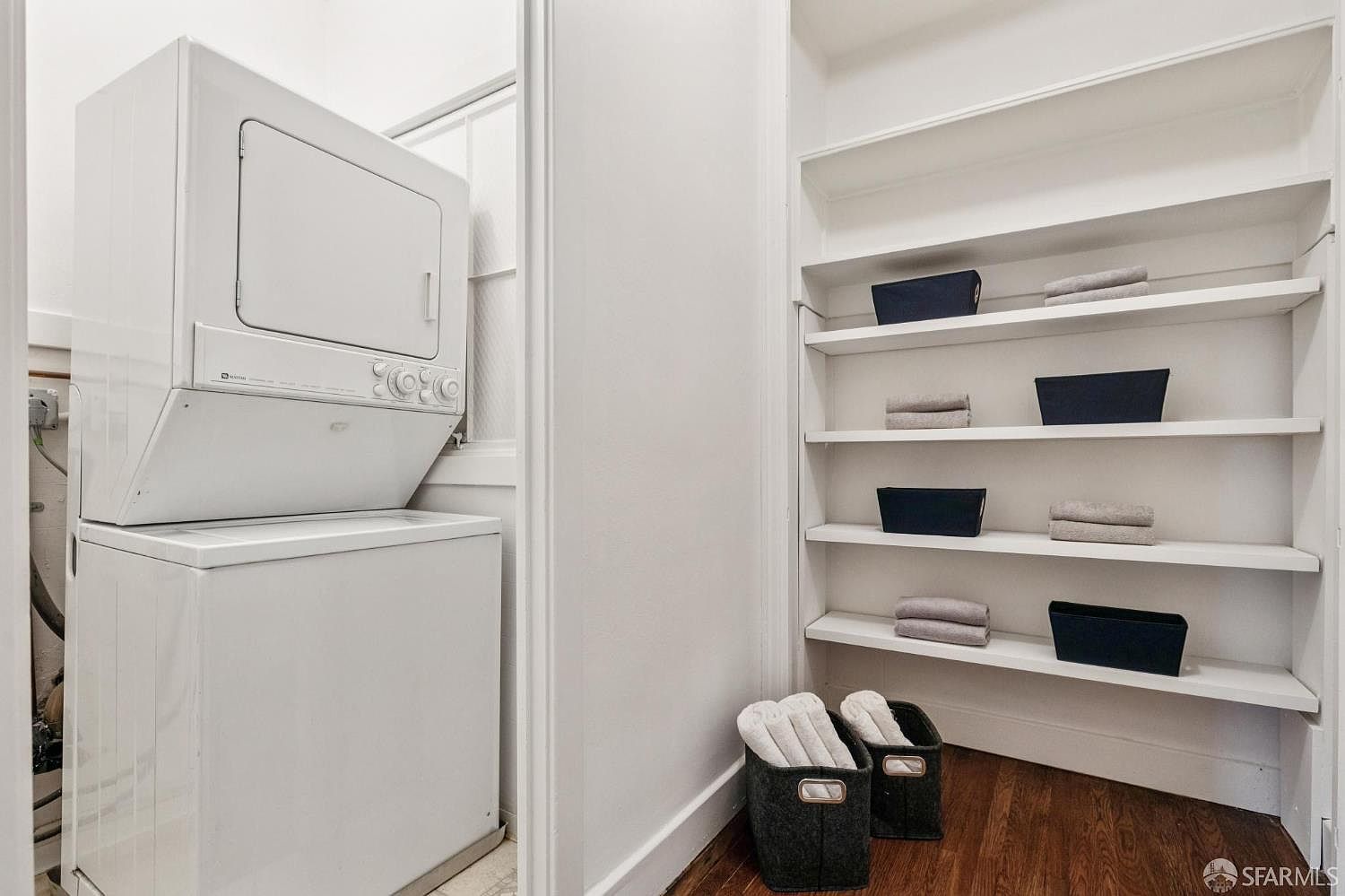 This is a well-organized laundry room featuring a stacked washer and dryer unit. Built-in shelving provides ample storage space, neatly arranged with dark storage bins and folded towels. The hardwood flooring adds a touch of warmth to the functional space.