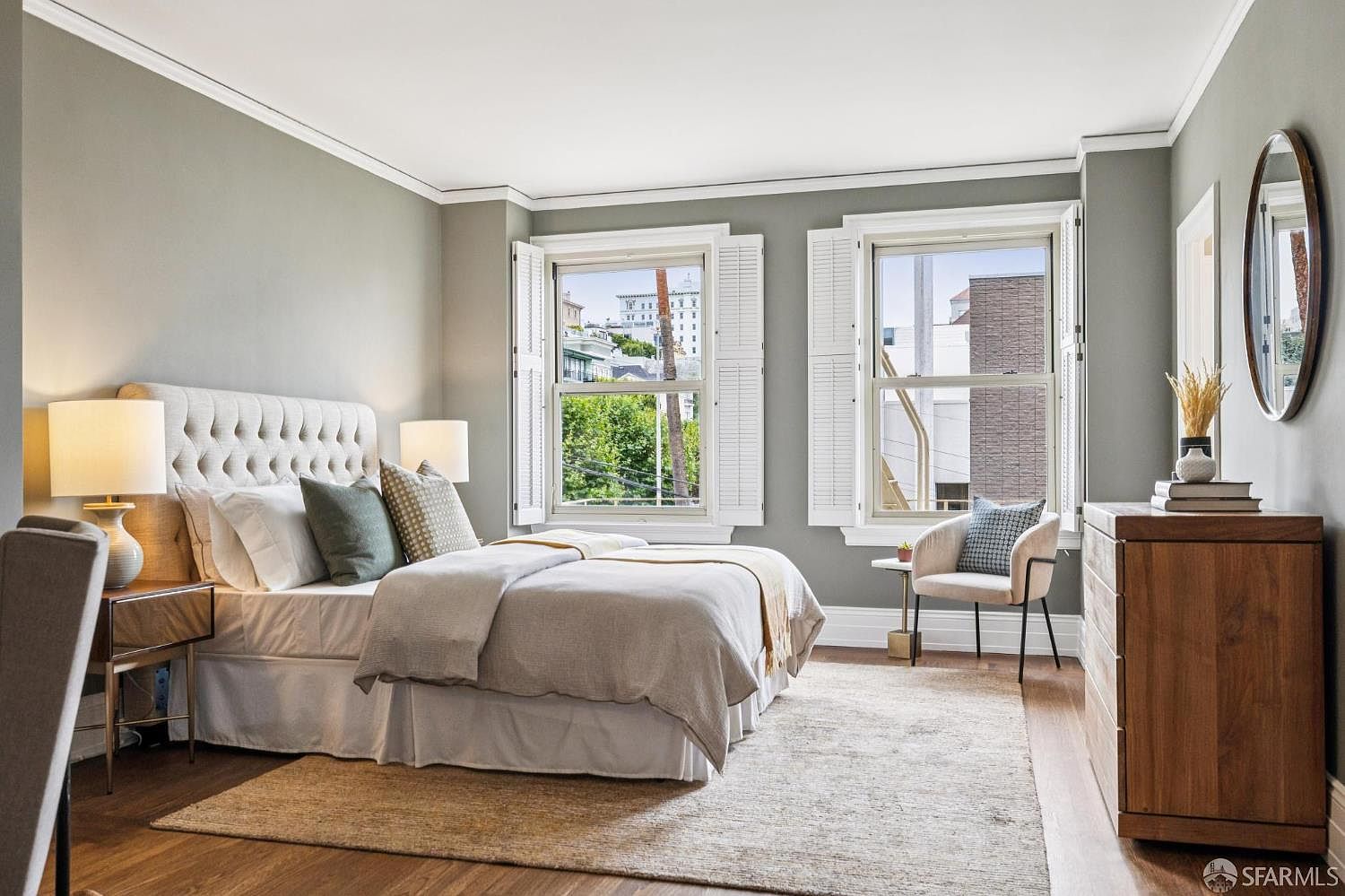 This is a well-lit primary bedroom featuring a large bed with a tufted headboard, neutral bedding, and decorative pillows. Two windows with white shutters provide natural light and views of the outside. A wooden dresser with a round mirror and a small chair add to the room's comfort and style, complemented by a soft area rug on hardwood floors.