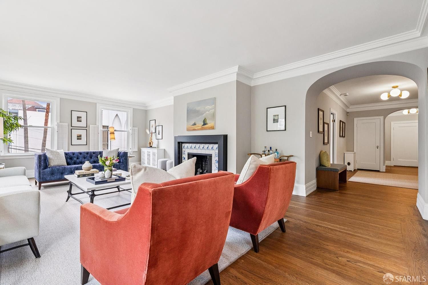 This is an interior shot of a living room featuring hardwood floors, a fireplace with decorative tiles, and an archway leading to another room. The room is furnished with a blue sofa, two orange armchairs, and a white coffee table. The walls are painted in a neutral gray tone, and the overall style is elegant and inviting.