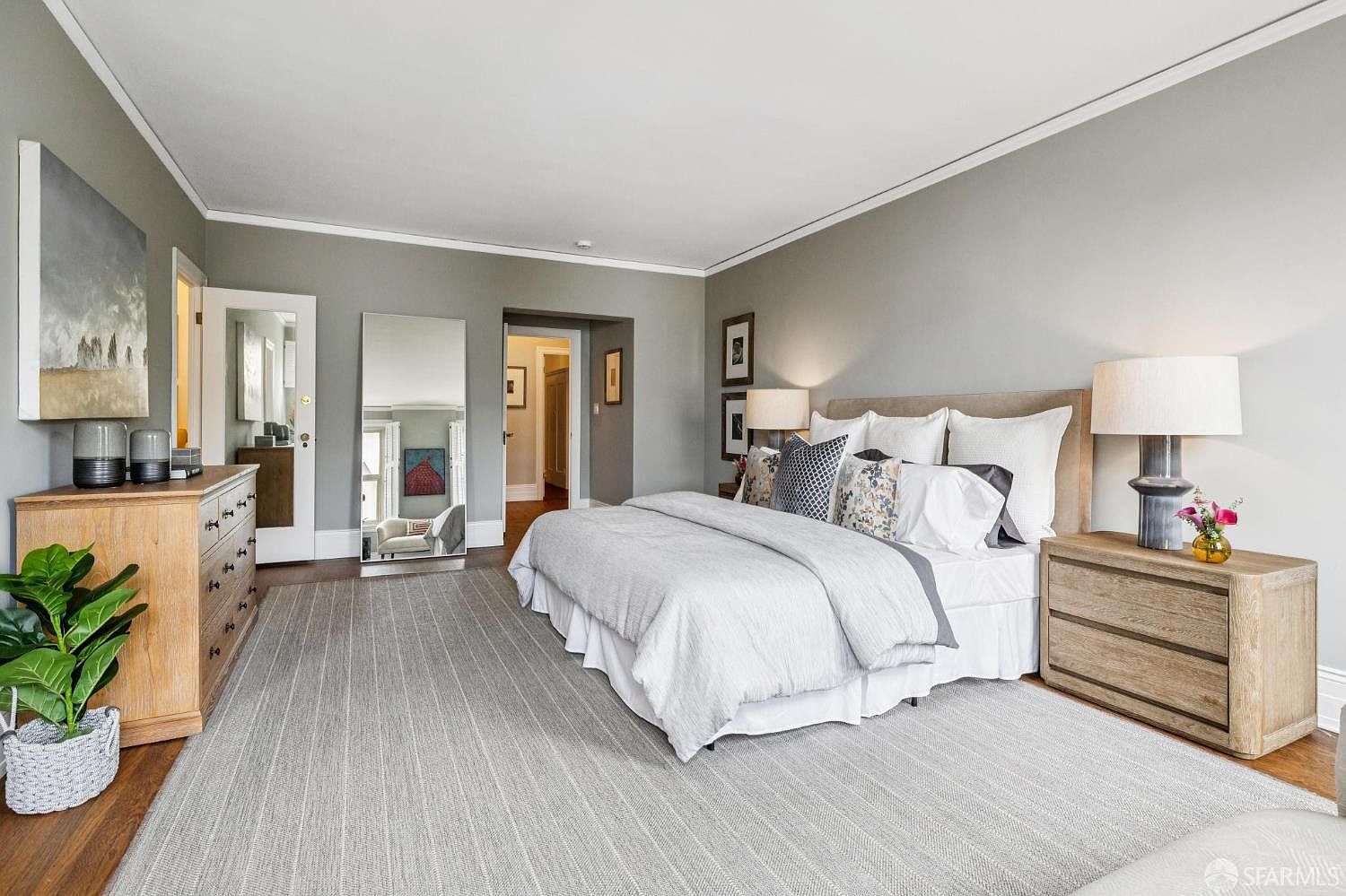 This is a primary bedroom featuring a neutral color palette with gray walls and a light gray carpet. A large bed with white linens and decorative pillows is the focal point, flanked by wooden nightstands and lamps. A wooden dresser with decorative vases and a large painting are visible on the left, and a full-length mirror adds depth to the room.