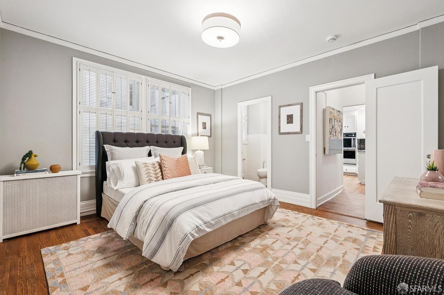 This is a well-lit primary bedroom featuring a queen-sized bed with a tufted headboard, striped bedding, and decorative pillows. The room has hardwood floors covered by an area rug with geometric patterns. Natural light streams in through shuttered windows, and there are doorways leading to other parts of the house, including a bathroom and possibly a kitchen.