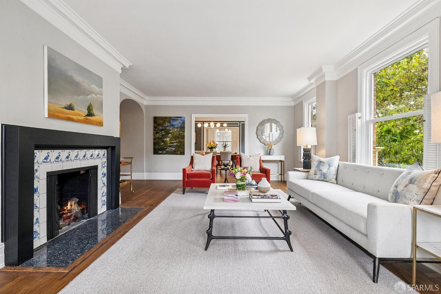 This is a bright and elegant living room featuring a fireplace with decorative tiles, a white sofa with patterned pillows, and two red armchairs. The room is well-lit with natural light from the windows and features hardwood floors partially covered by a light-colored area rug. The overall impression is one of sophistication and comfort.