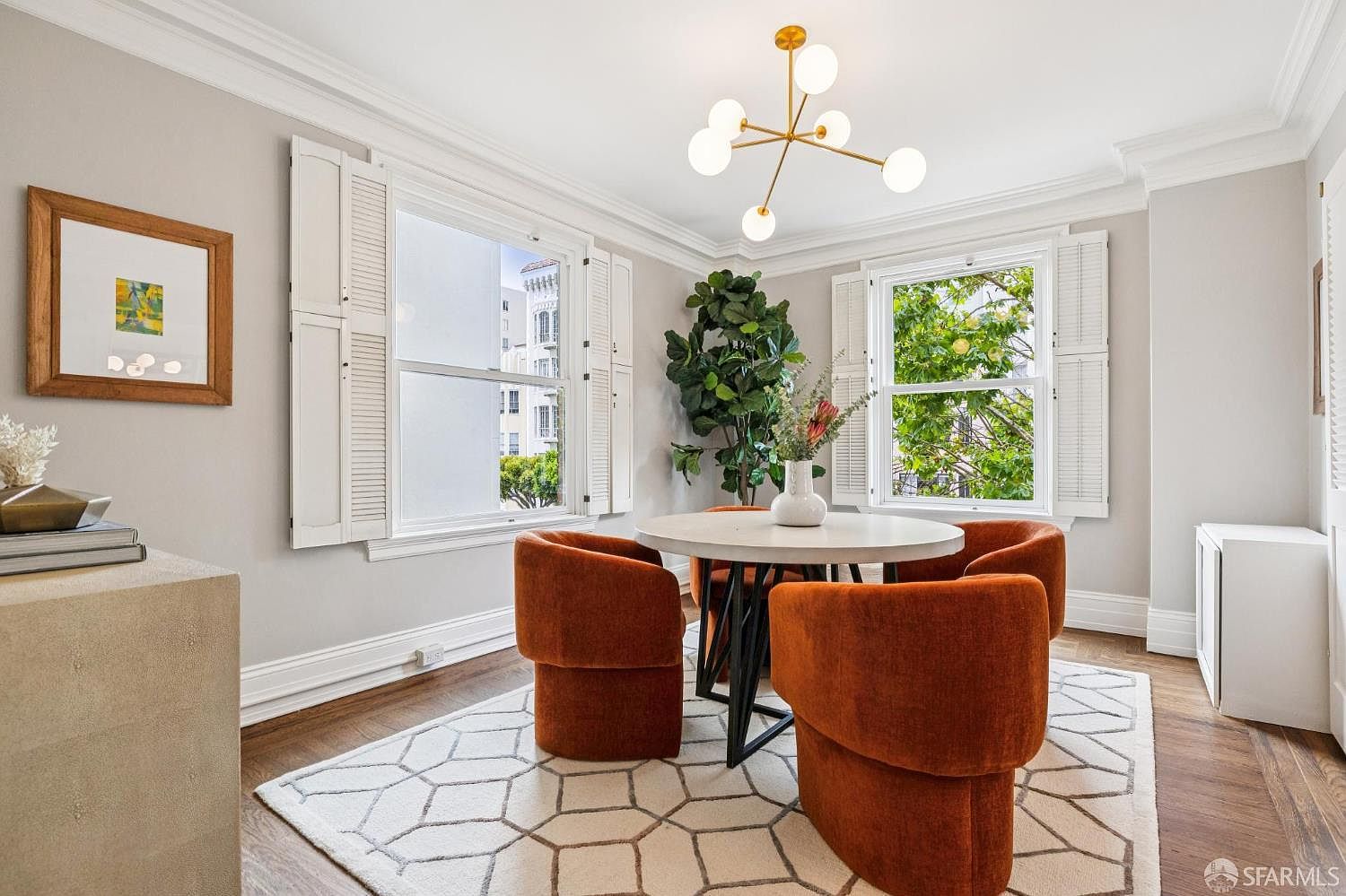 This is an interior shot of a dining room featuring a round table with four orange upholstered chairs. The room is well-lit with natural light coming through two windows with white shutters. A modern chandelier hangs above the table, and a patterned rug covers the hardwood floor, creating a cozy and stylish dining space.