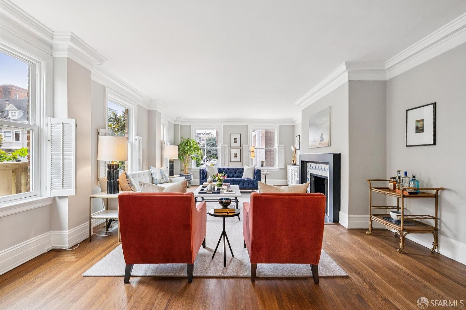 This is a well-lit living room featuring hardwood floors, a fireplace, and large windows that provide ample natural light. The room is furnished with two orange armchairs, a blue sofa, and a bar cart, creating a comfortable and inviting atmosphere. The perspective is from the entrance of the room, showcasing the depth and arrangement of the furniture.