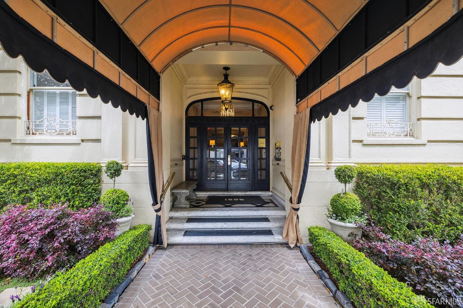 The image showcases an elegant entryway to a building, featuring a covered entrance with a black and beige awning and matching curtains. The entrance is framed by manicured greenery and a brick walkway, leading to a set of glass doors. A vintage-style lantern hangs above the doorway, adding to the classic and sophisticated ambiance.