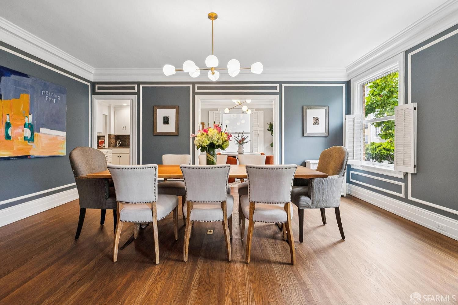 This is an interior shot of a dining room featuring a wooden dining table surrounded by upholstered chairs. The room is decorated with framed artwork and a modern chandelier, with natural light coming in from a window with shutters. The walls are painted a dark gray with white trim, creating a sophisticated and inviting atmosphere.