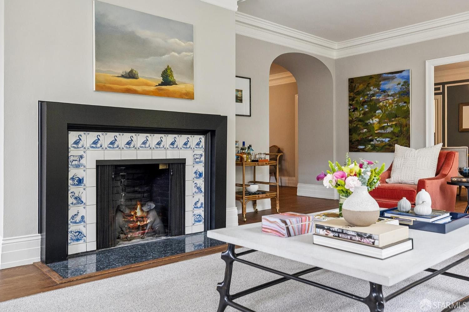 This is an interior shot of a living room featuring a fireplace with blue and white tiles and a black frame. Above the fireplace hangs a landscape painting. The room also includes a coffee table with books and decorative items, a red armchair, and an arched doorway leading to another room. The overall style is elegant and cozy.