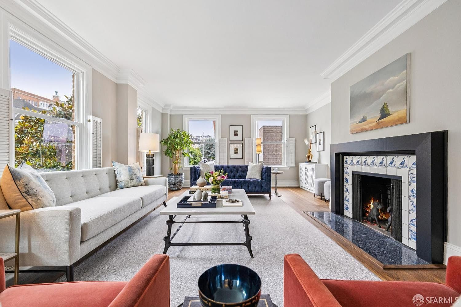 This is a well-lit living room featuring a light gray sofa, a blue velvet sofa, and two orange armchairs. A fireplace with decorative tiles adds a focal point, and large windows provide ample natural light. The room is decorated with plants and artwork, creating a comfortable and inviting atmosphere.