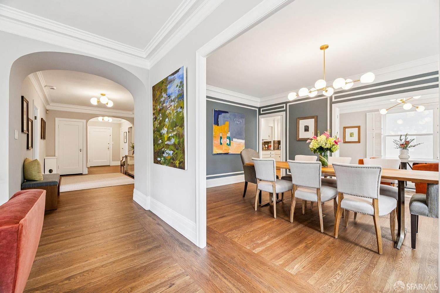This image showcases an elegant dining room adjacent to a hallway, featuring hardwood floors laid in a herringbone pattern. The dining area is furnished with a wooden table surrounded by upholstered chairs, illuminated by a modern chandelier. The walls are painted in a sophisticated gray tone with white trim, and artwork adorns the walls, adding a touch of color and personality to the space.