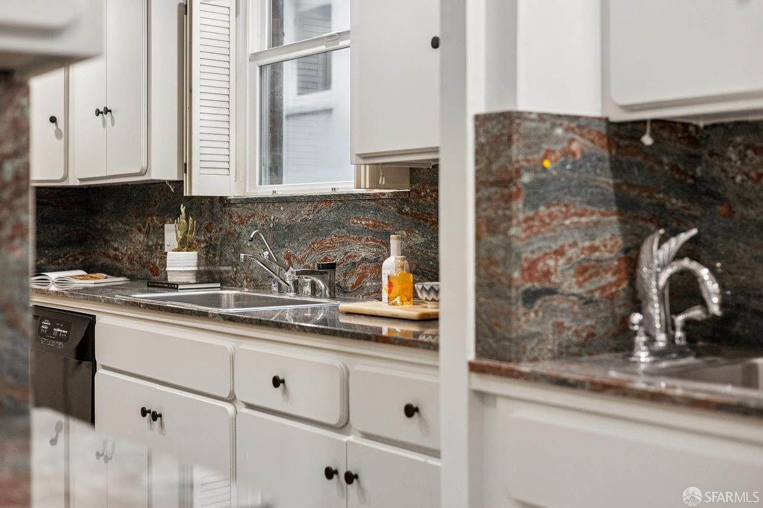 This kitchen features white cabinetry with black hardware, a dark granite countertop and backsplash, and stainless steel appliances. A window above the sink provides natural light, and decorative items add a touch of personality. The overall impression is a clean and functional space with a classic design.
