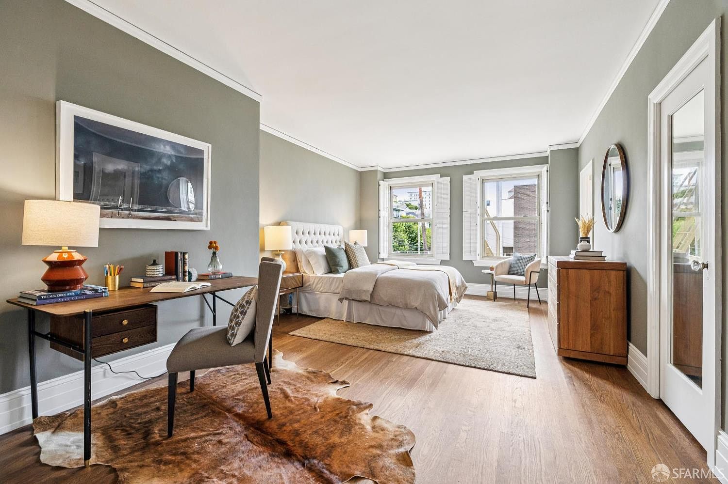 This is a well-lit primary bedroom featuring hardwood floors, a neutral color palette, and a blend of modern and traditional elements. The room includes a tufted headboard bed, a desk area with a modern lamp, and a dresser with a round mirror. Natural light streams in through the windows, creating a warm and inviting atmosphere.