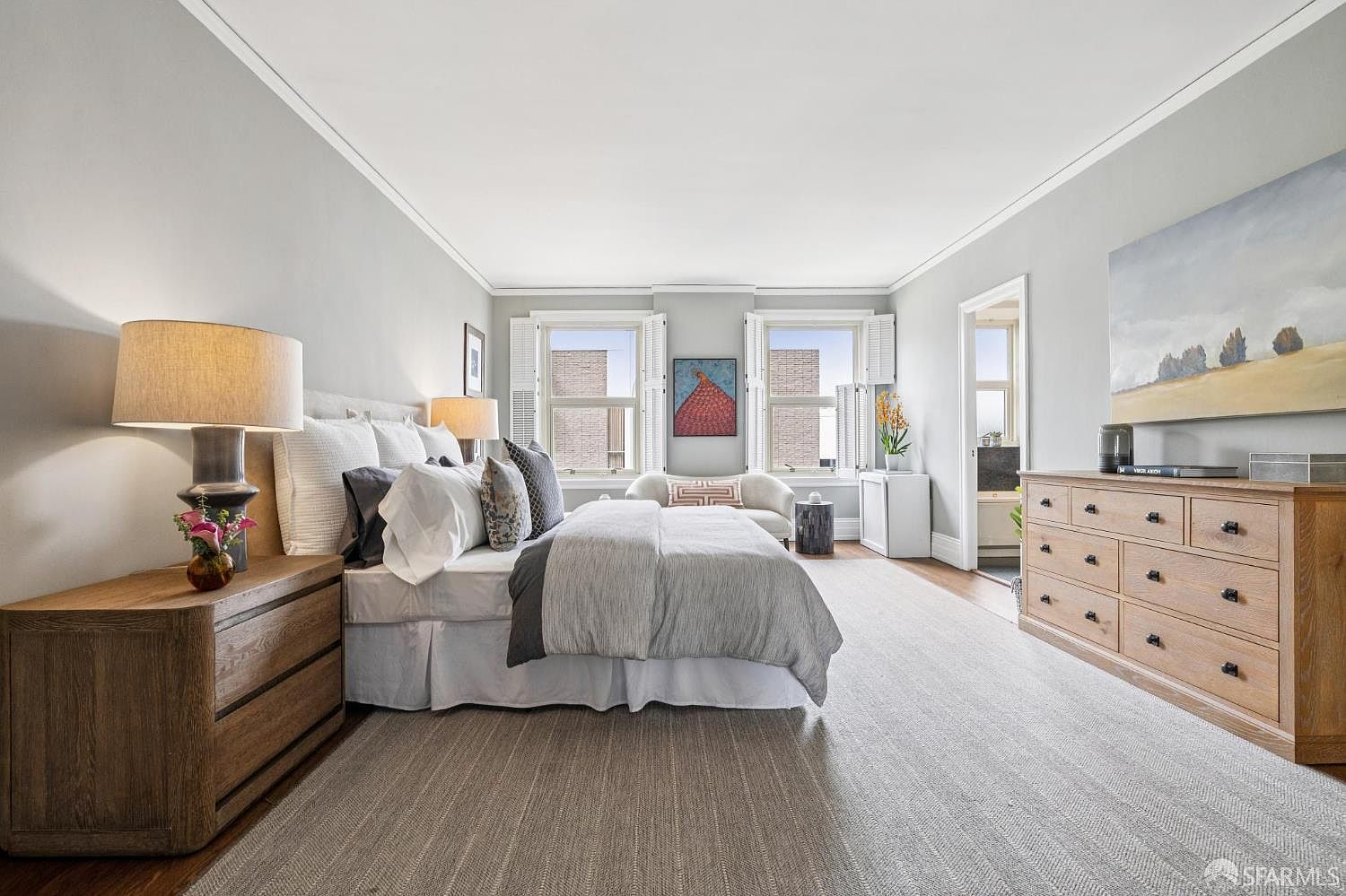 This is a well-lit primary bedroom featuring a large bed with multiple pillows and a gray duvet. The room is decorated in neutral tones with light gray walls and a beige carpet. There are two windows with white shutters, a wooden dresser, and a nightstand with a lamp, creating a serene and inviting atmosphere.
