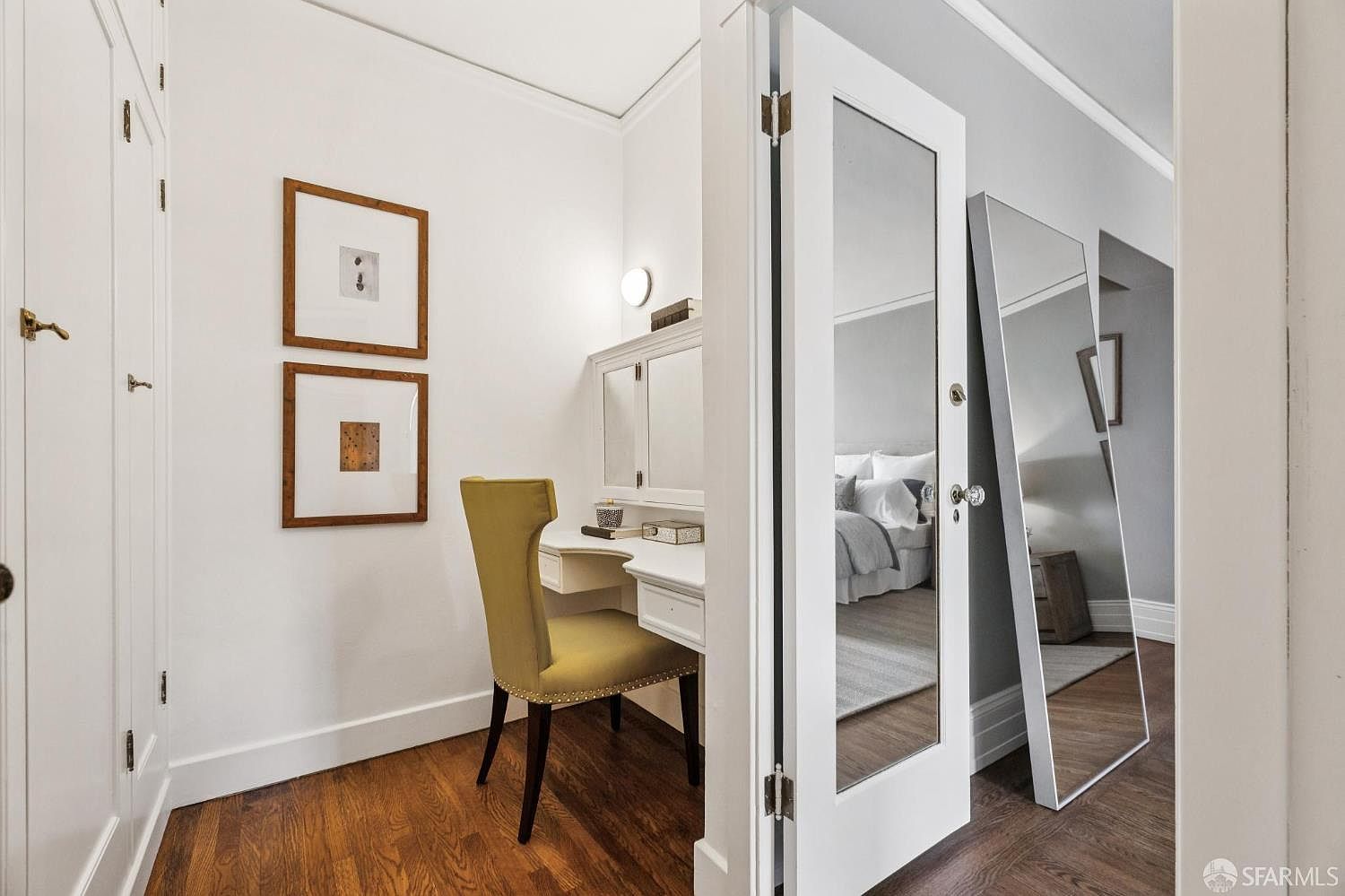 This interior shot showcases a charming bedroom nook featuring a built-in vanity with a yellow chair, complemented by framed artwork on the wall. The room is partially visible through mirrored closet doors, offering a glimpse of the bed and further enhancing the sense of space. The hardwood floors and white trim contribute to a clean and inviting aesthetic.
