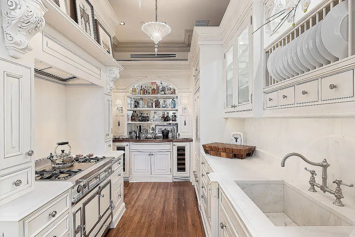 This elegant, high-end kitchen features classic white cabinetry, a professional-grade range with a decorative hood, and a deep farmhouse-style sink set into a stone countertop. The space is accented by ornate architectural moldings, a glass-fronted dish rack, and a dedicated bar area with open shelving at the far end. The perspective is a straight-on, eye-level shot looking down the galley-style layout, emphasizing the sophisticated craftsmanship and bright, airy atmosphere.