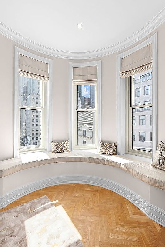 This elegant interior space features a curved window nook with a built-in bench seat, offering a panoramic view of the city skyline. The room is finished with light-toned walls, intricate crown molding, and warm-toned herringbone wood flooring. The bright, airy atmosphere is enhanced by the natural light streaming through the three tall windows, which are fitted with neutral-colored roman shades.