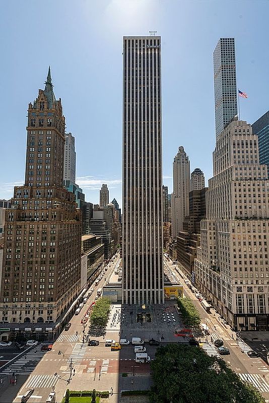 This high-angle aerial view captures a prominent, slender skyscraper in the heart of a dense urban environment, flanked by historic and modern high-rise buildings. The perspective looks down onto a bustling city street intersection and a public plaza at the base of the central tower, which features a distinct glass-enclosed entrance. The scene is bathed in bright daylight, emphasizing the architectural contrast between the classic masonry facades of the surrounding structures and the sleek, vertical lines of the central skyscraper.