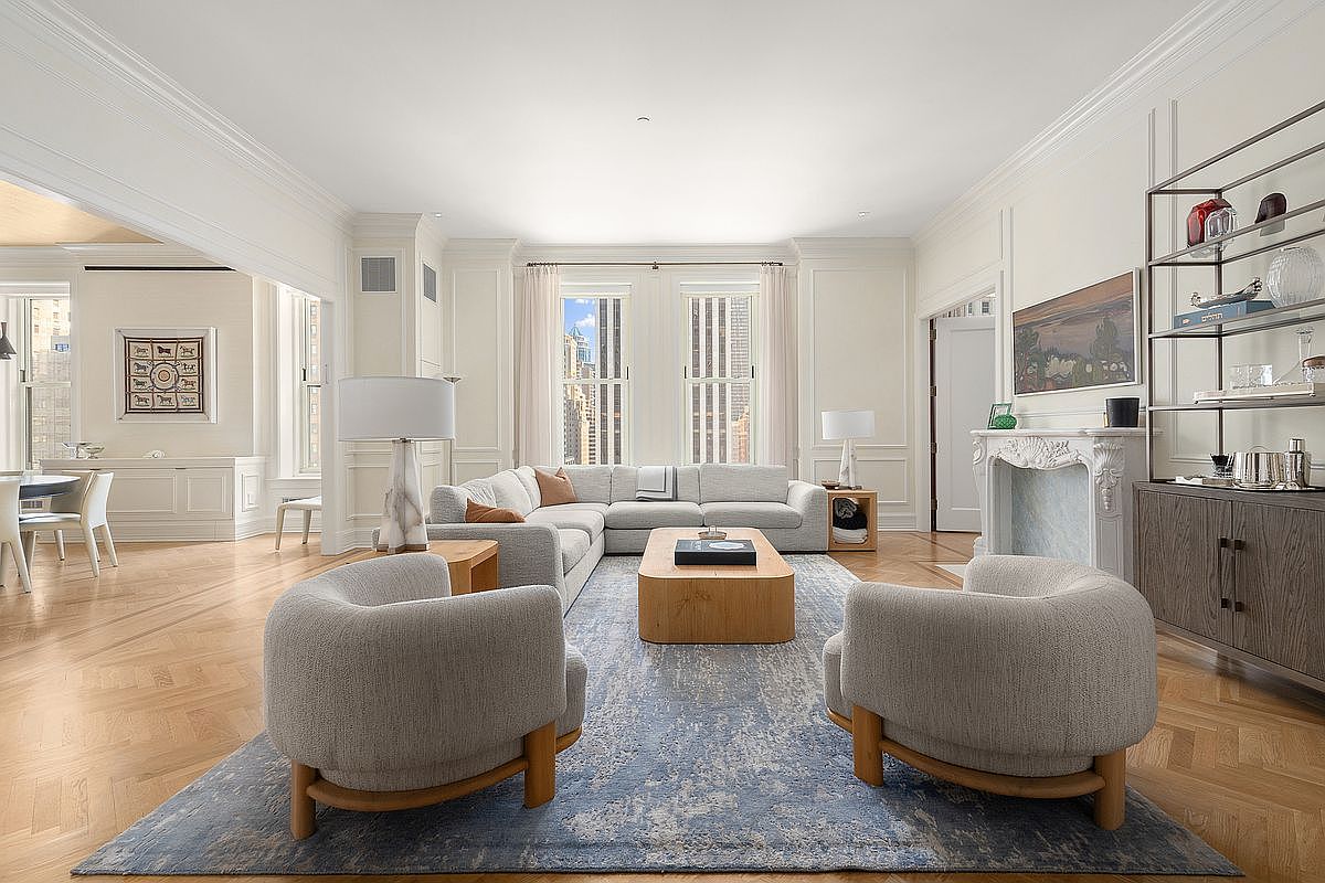 This elegant living room features a spacious, light-filled layout with classic architectural molding and polished herringbone wood floors. The room is anchored by a large, neutral-toned sectional sofa and two curved armchairs arranged around a low, light wood coffee table on a textured blue area rug. A decorative fireplace with a carved mantel serves as a focal point, complemented by a tall metal shelving unit and a view of city buildings through large, symmetrical windows.