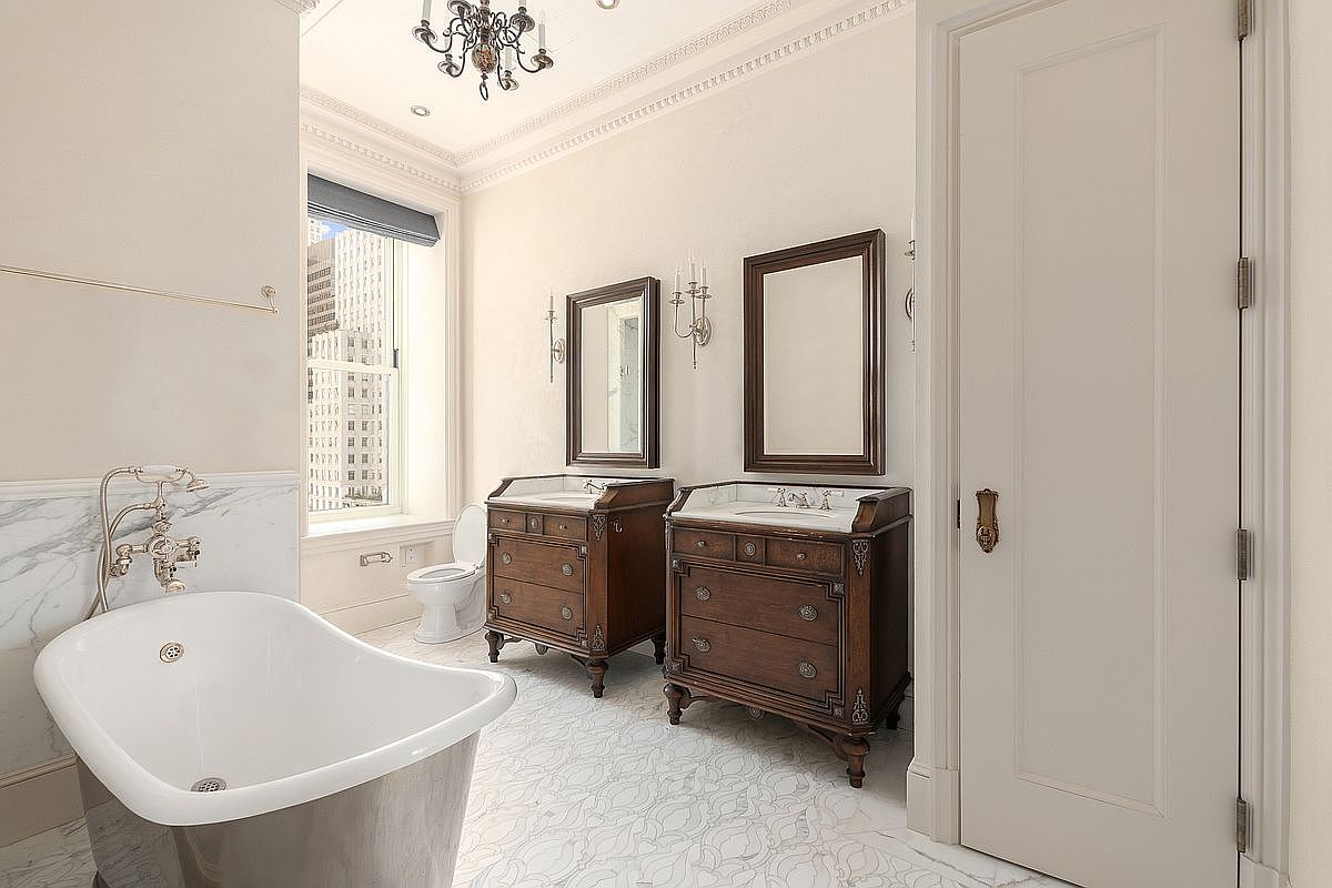 This elegant primary bathroom features a classic freestanding soaking tub with polished metal fixtures and a dual-vanity setup with dark wood cabinetry and stone countertops. The space is illuminated by a decorative chandelier and wall-mounted sconces, while a large window provides natural light and a view of the city skyline. The room is finished with intricate patterned tile flooring and crisp white walls, creating a sophisticated and luxurious atmosphere.