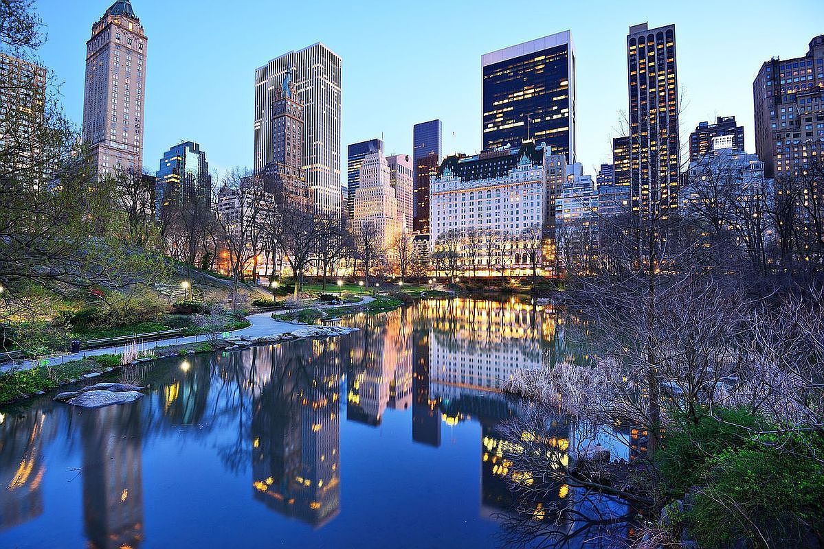 This scenic view captures the tranquil pond in Central Park, reflecting the iconic New York City skyline at dusk. The composition highlights a lush, tree-lined foreground leading to a dense cluster of historic and modern skyscrapers, creating a striking contrast between nature and urban architecture. The soft, ambient lighting of the city buildings creates a serene and prestigious atmosphere, emphasizing the prime location of the surrounding real estate.