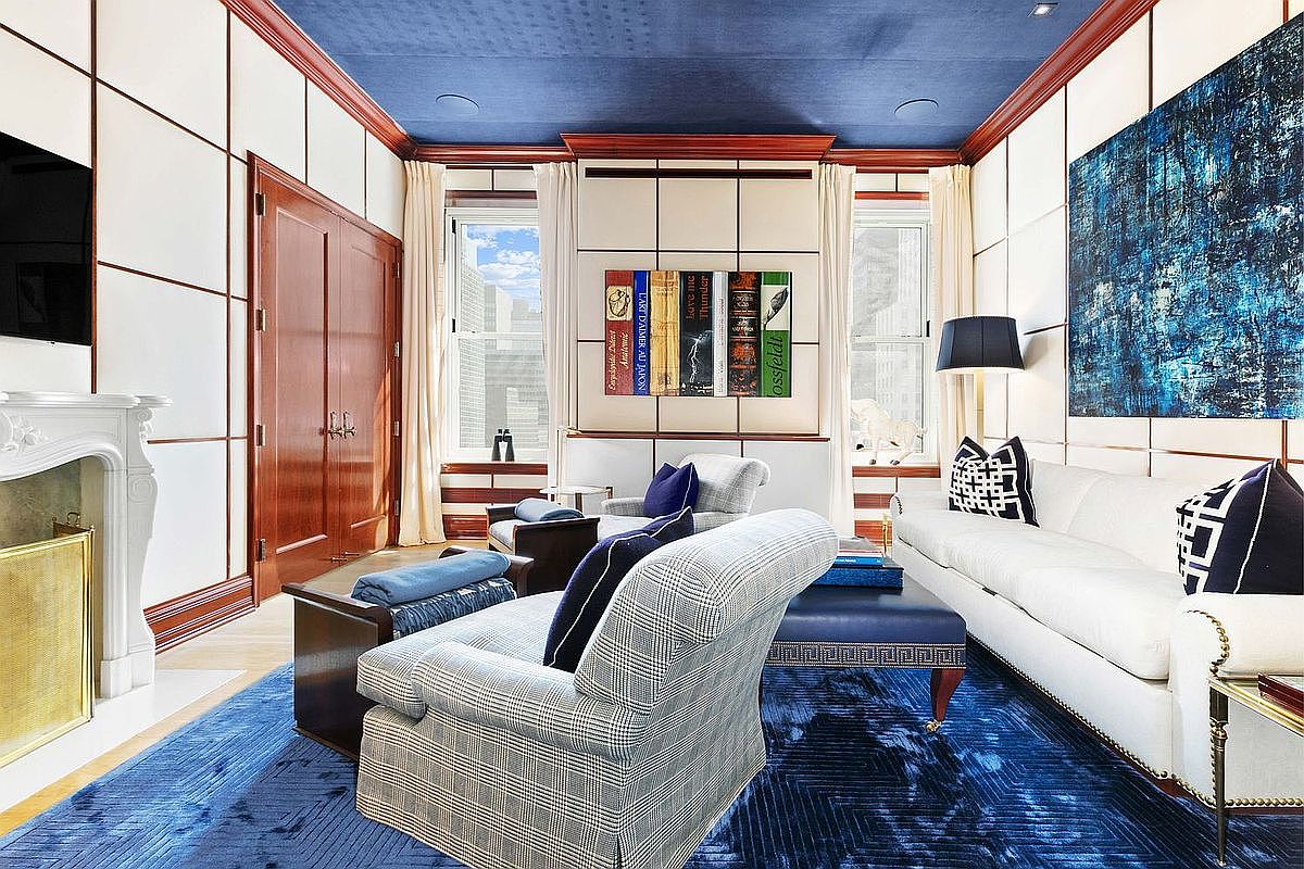 This sophisticated living room features a bold, deep blue ceiling and a matching area rug that anchors the space. The room is appointed with a white sofa, patterned armchairs, and a central ottoman, all set against walls with a grid-paneled design. A fireplace with a classic mantel sits to the left, while large windows draped in light curtains allow natural light to illuminate the room's elegant, high-contrast aesthetic.