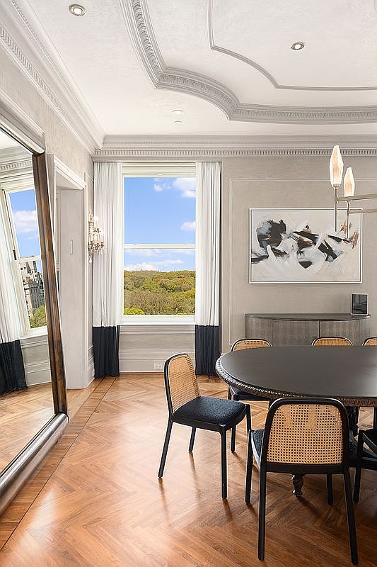 This elegant dining room features rich herringbone-patterned wood flooring and intricate crown molding that highlights the sophisticated architectural detail. A round dark-toned table is surrounded by modern chairs with woven cane backs, positioned near a large window that frames a scenic view of lush greenery. The space is accented by a contemporary chandelier, a large floor mirror, and a piece of abstract wall art, creating a refined and airy atmosphere.