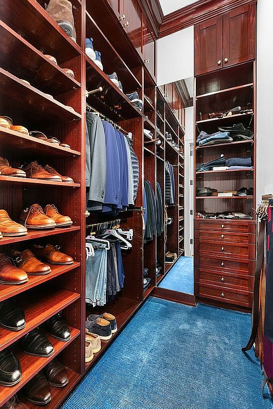This spacious walk-in closet features extensive custom dark wood cabinetry with integrated shelving, hanging rods, and a bank of drawers. The room is accented by a large mirror that enhances the sense of depth, while a vibrant blue carpet provides a bold contrast to the rich wood tones. The perspective is a straight-on view looking down the narrow aisle, showcasing the organized storage capacity of the space.