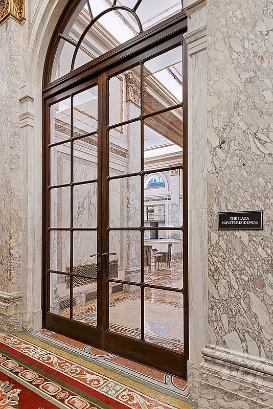 This elegant entryway features a set of tall, multi-paned glass doors framed in dark wood, set within a grand archway. The surrounding walls are clad in polished, light-colored stone with intricate veining, and a sign identifies the area as 'The Plaza Private Residences.' A vibrant, patterned carpet leads up to the threshold, creating a luxurious and formal transition between spaces.