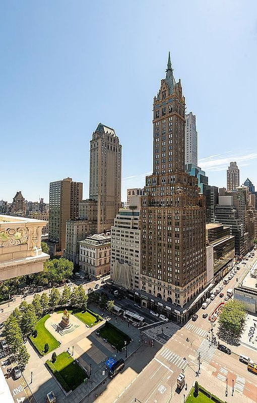This high-angle aerial perspective captures a prominent, ornate skyscraper with a distinctive Gothic-inspired spire, standing as a focal point among the dense urban architecture of New York City. Below, a manicured public park with green lawns and pathways provides a serene contrast to the bustling city streets and traffic visible at the intersection. The clear blue sky and bright daylight emphasize the intricate masonry and verticality of the surrounding high-rise buildings.