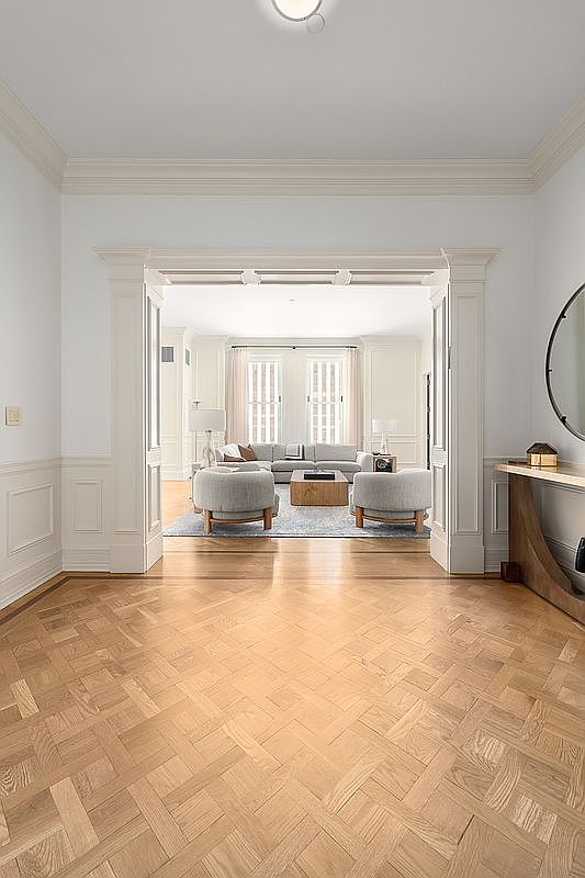 This elegant entryway features a sophisticated basket-weave patterned light wood floor that leads the eye through a grand, cased opening into a bright living area. The space is defined by classic architectural details, including wainscoting and crown molding, finished in a crisp, neutral palette. A sleek console table sits to the right, while the open-concept transition creates a seamless flow between the foyer and the sun-drenched living room beyond.