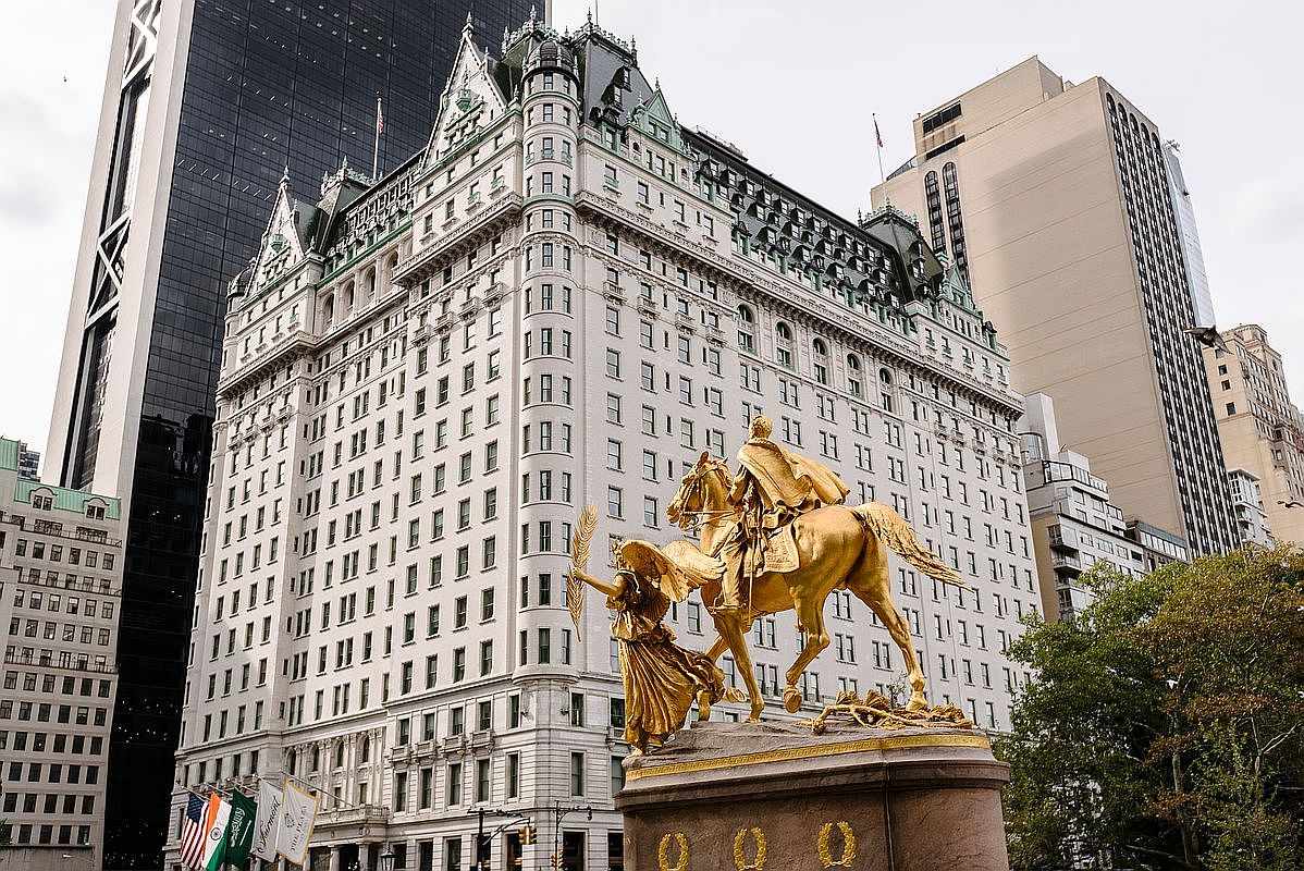 This iconic view captures the historic Plaza Hotel in New York City, showcasing its grand French Renaissance-inspired architecture and ornate detailing. In the foreground, the golden General William Tecumseh Sherman Monument stands prominently against the backdrop of the hotel's white facade and steep, dark-toned roof. The perspective is cinematic and low-angle, emphasizing the scale of the landmark building alongside the surrounding urban skyline.